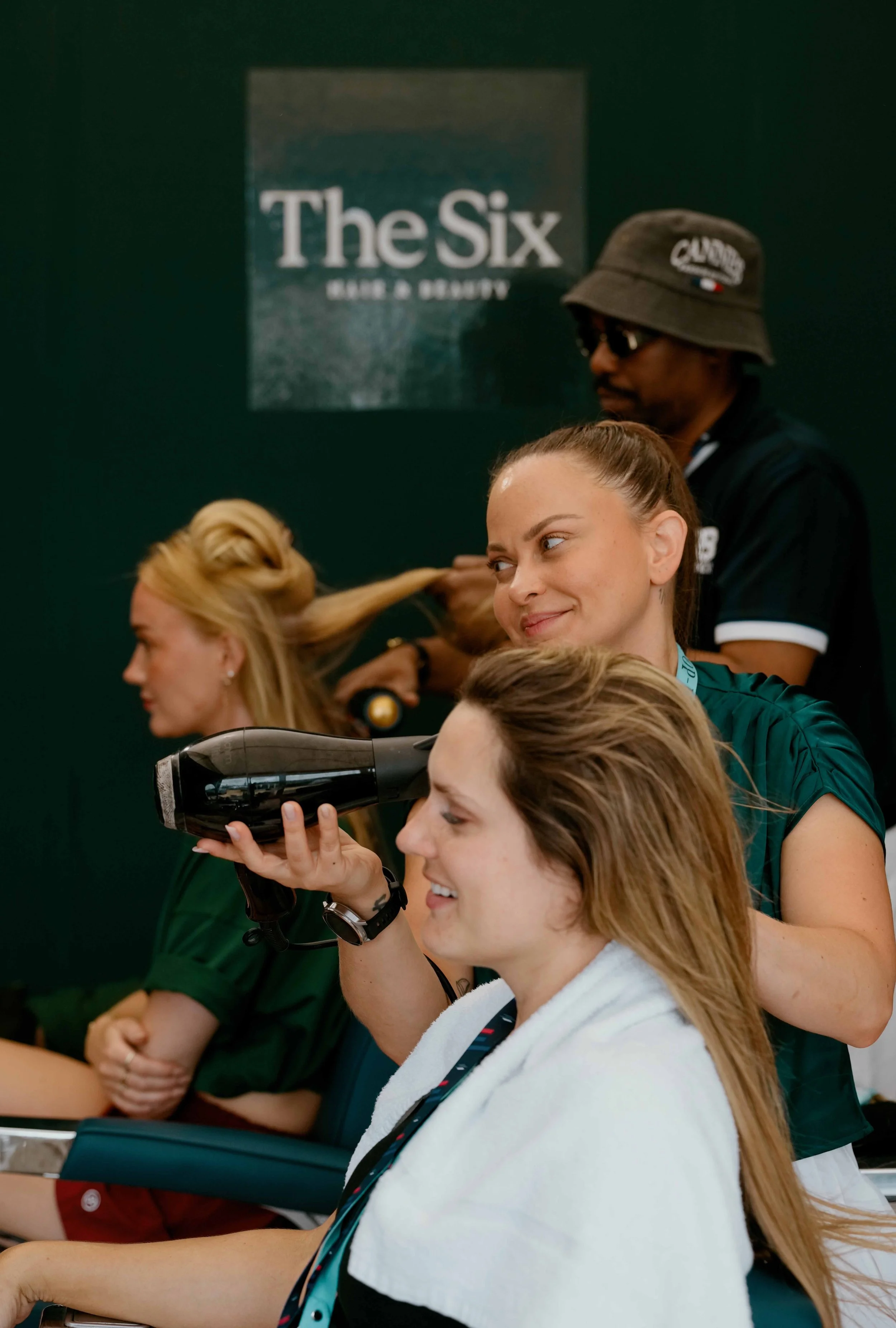 Women sitting in a row, including a blonde woman smiling, a woman with a towel over her shoulders, and a woman holding a hairdryer, with a man in a bucket hat styling hair in the background near a sign that reads "The Six."