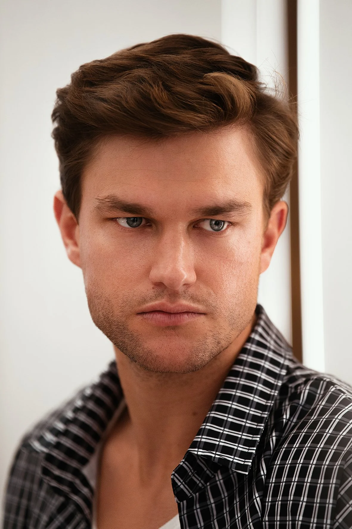 Close-up of a young man with brown hair, wearing a black and white checkered shirt, looking slightly to his left with a serious expression.
