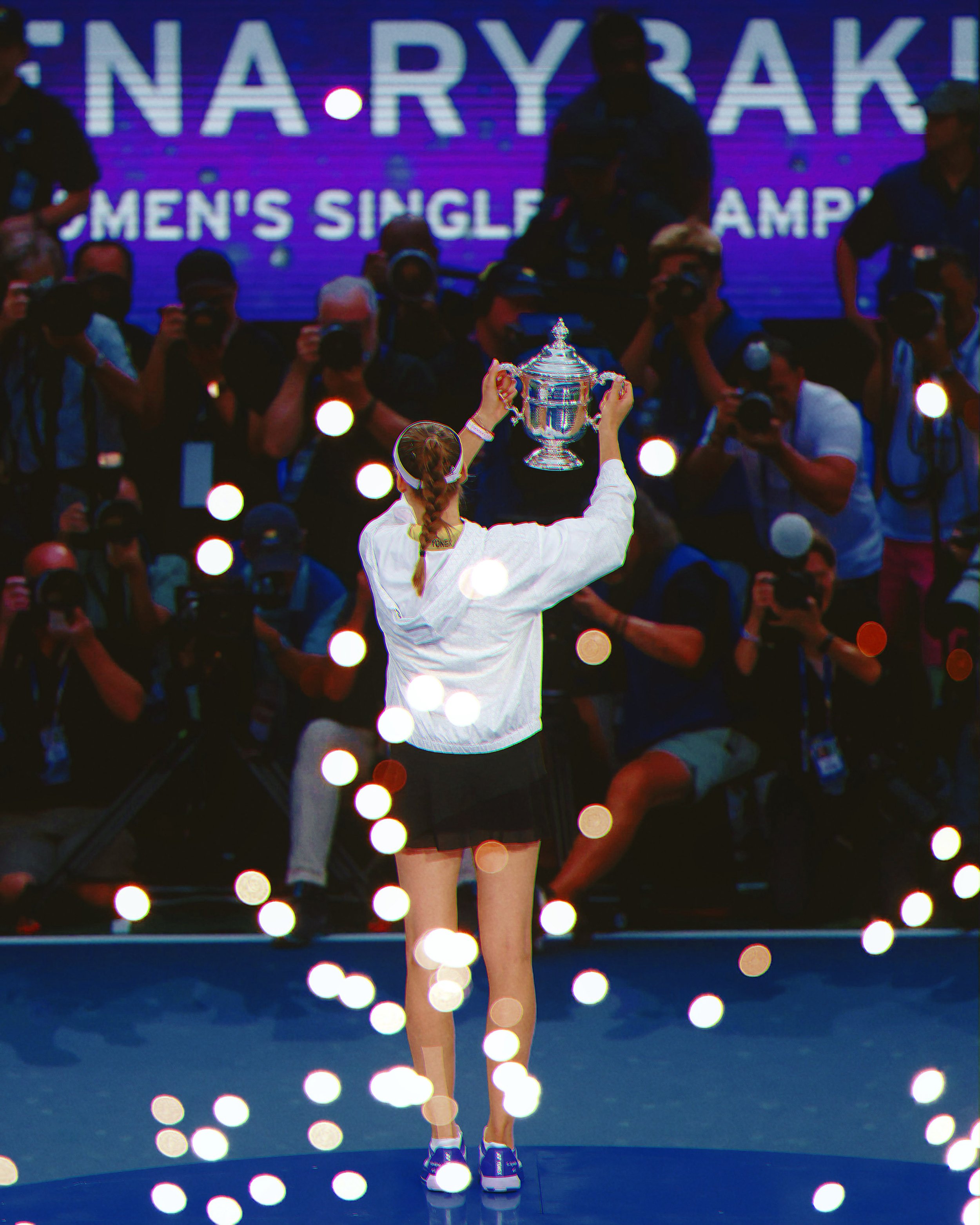 A female golfer with a braid holding a large silver trophy on a stage with a sign in the background that reads 'N/A RY '. Photographers are taking pictures of her.