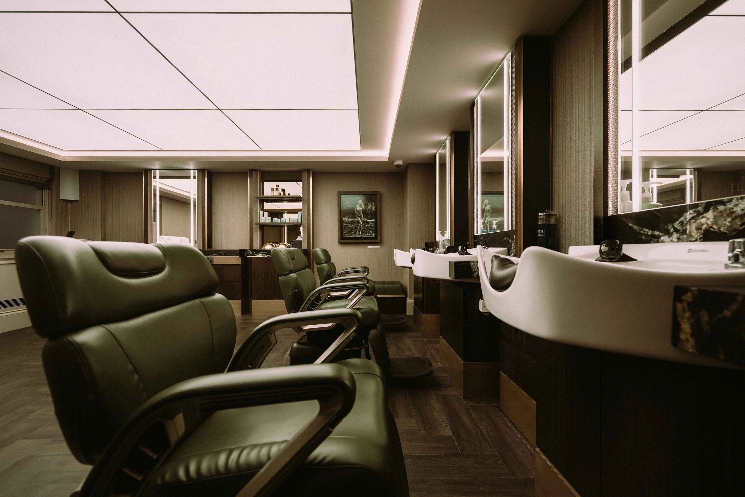Barbering area at The Six, Mayfair, featuring leather chairs, a marble countertop sink, and wall-mounted mirrors with a framed photograph and shelves in the background.
