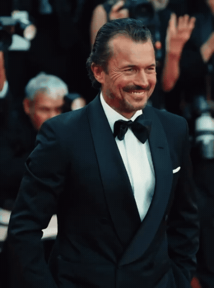 A man smiling in a tuxedo at a formal event, with photographers and other attendees in the background.