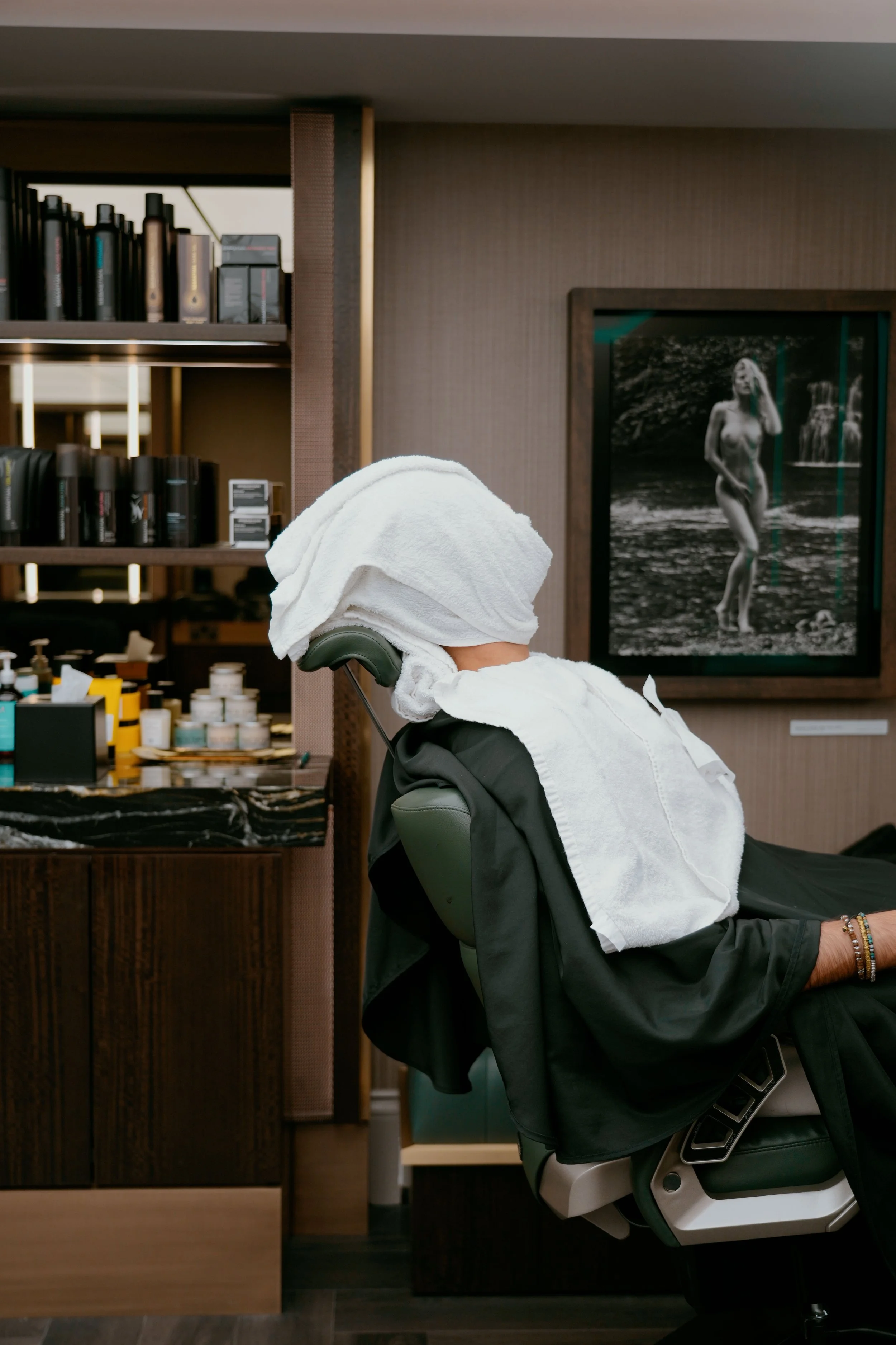 A person lying on a barber chair with their head and face wrapped in a white hot towel.