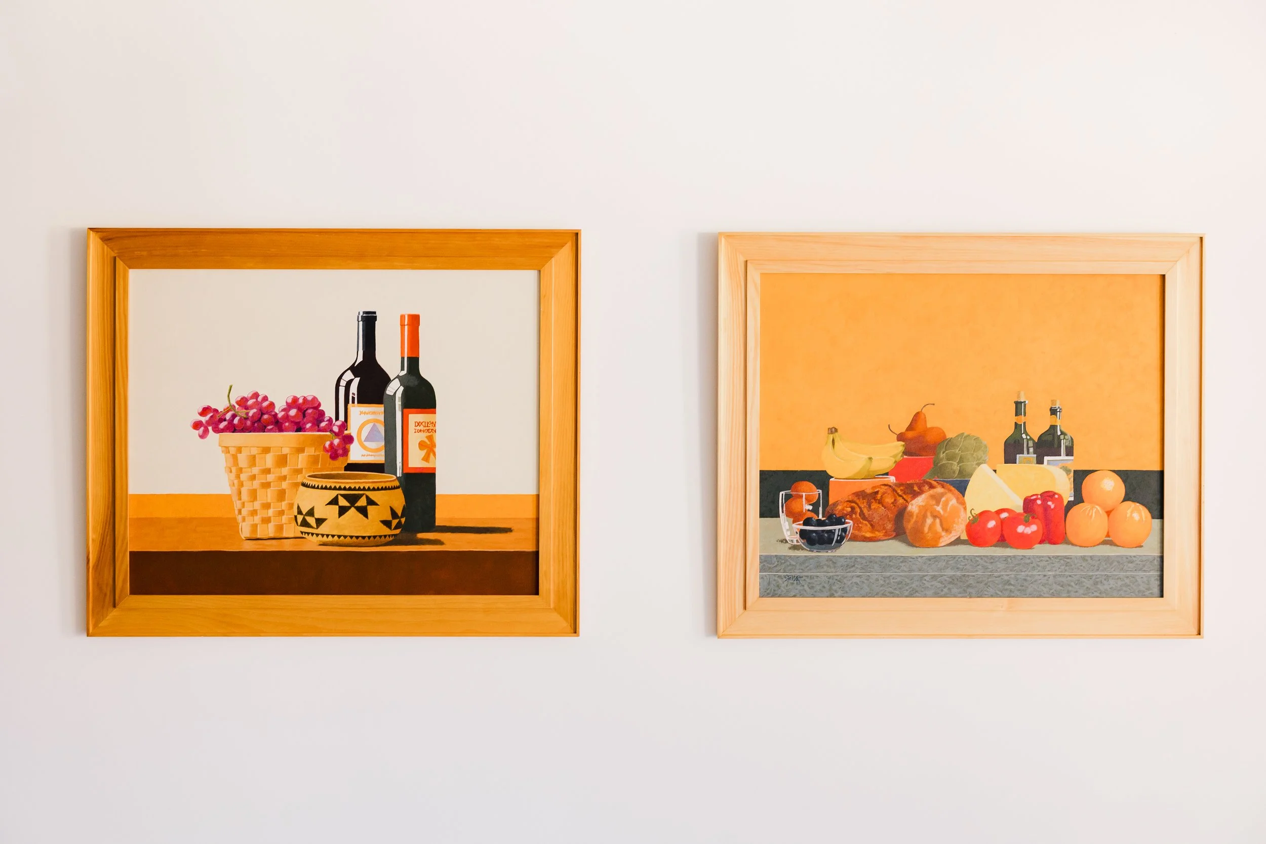 Two framed still life paintings hanging on a white wall, one with grapes, bottles, and a basket; the other with vegetables and fruits.