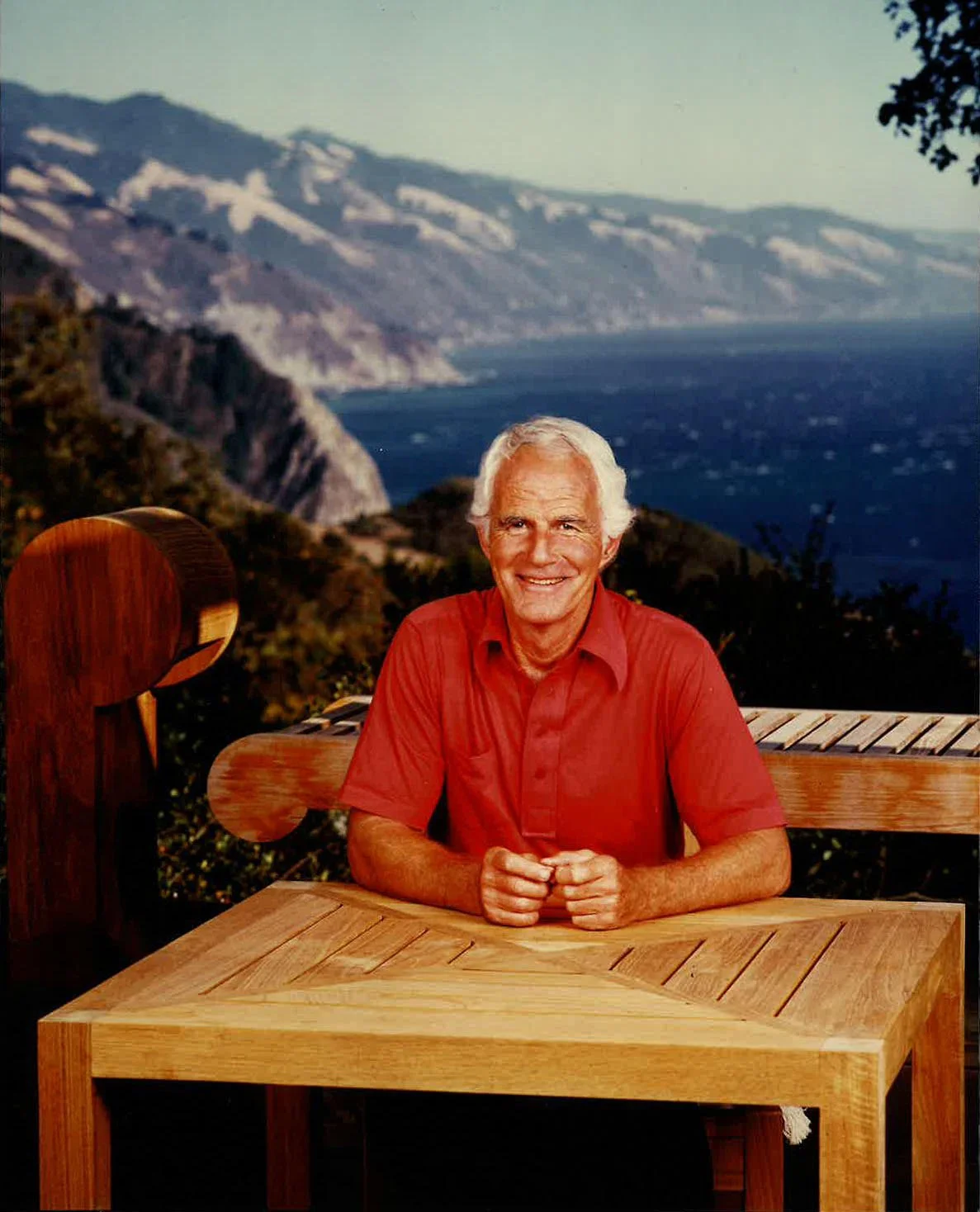 An older man with white hair, wearing a red shirt, sitting at a wooden table outdoors with a landscape of cliffs, water, and mountains in the background.