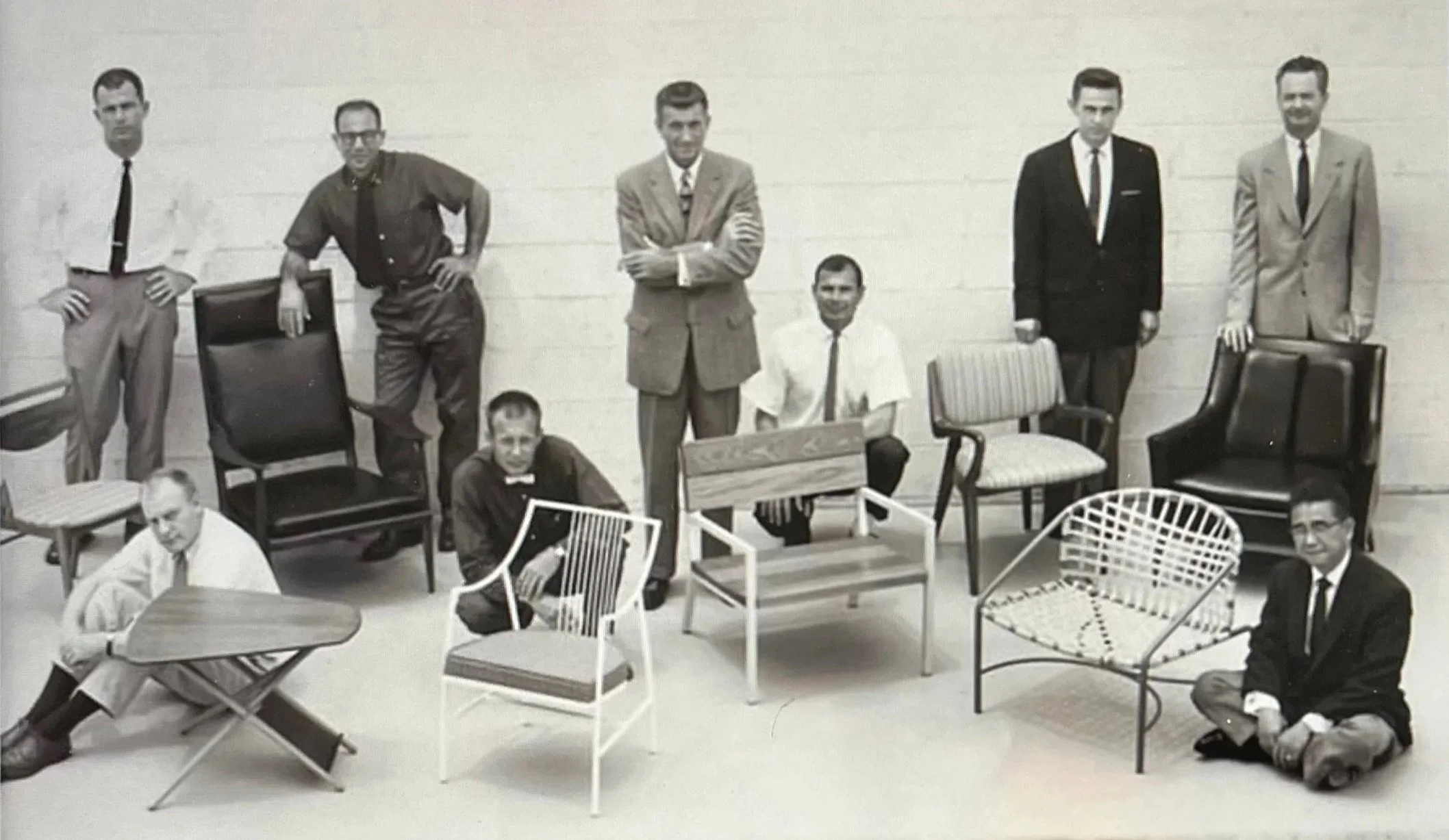 Black and white photo of nine renowned furniture designers posing and sitting among various chairs and furniture in a room with a plain wall.