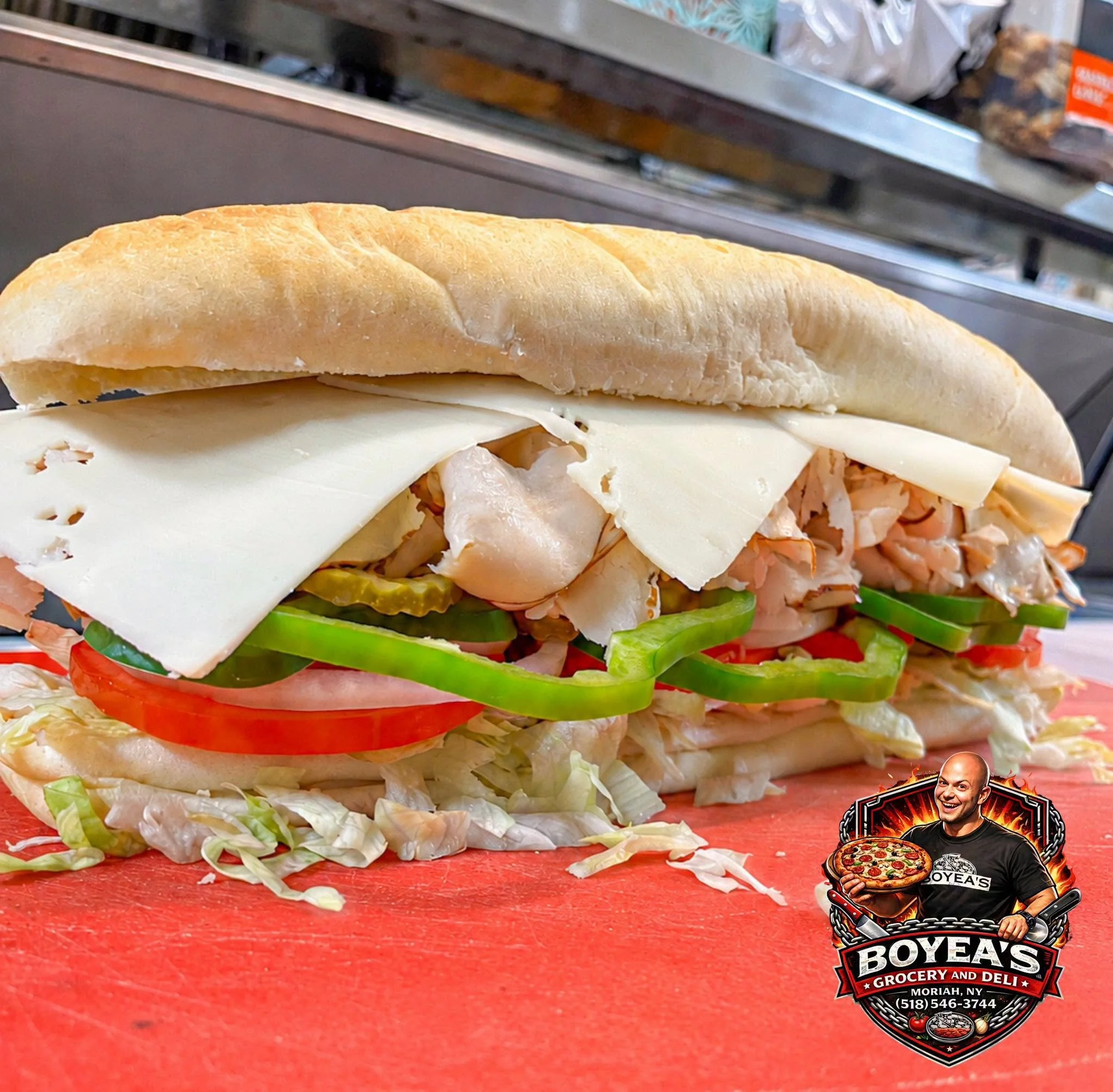 A close-up of a submarine sandwich with lettuce, tomato, pickles, turkey, cheese, and a hoagie roll on a red surface at Boyeas Grocery and Deli in Moriah, NY. The background shows a grocery store aisle with shelves.