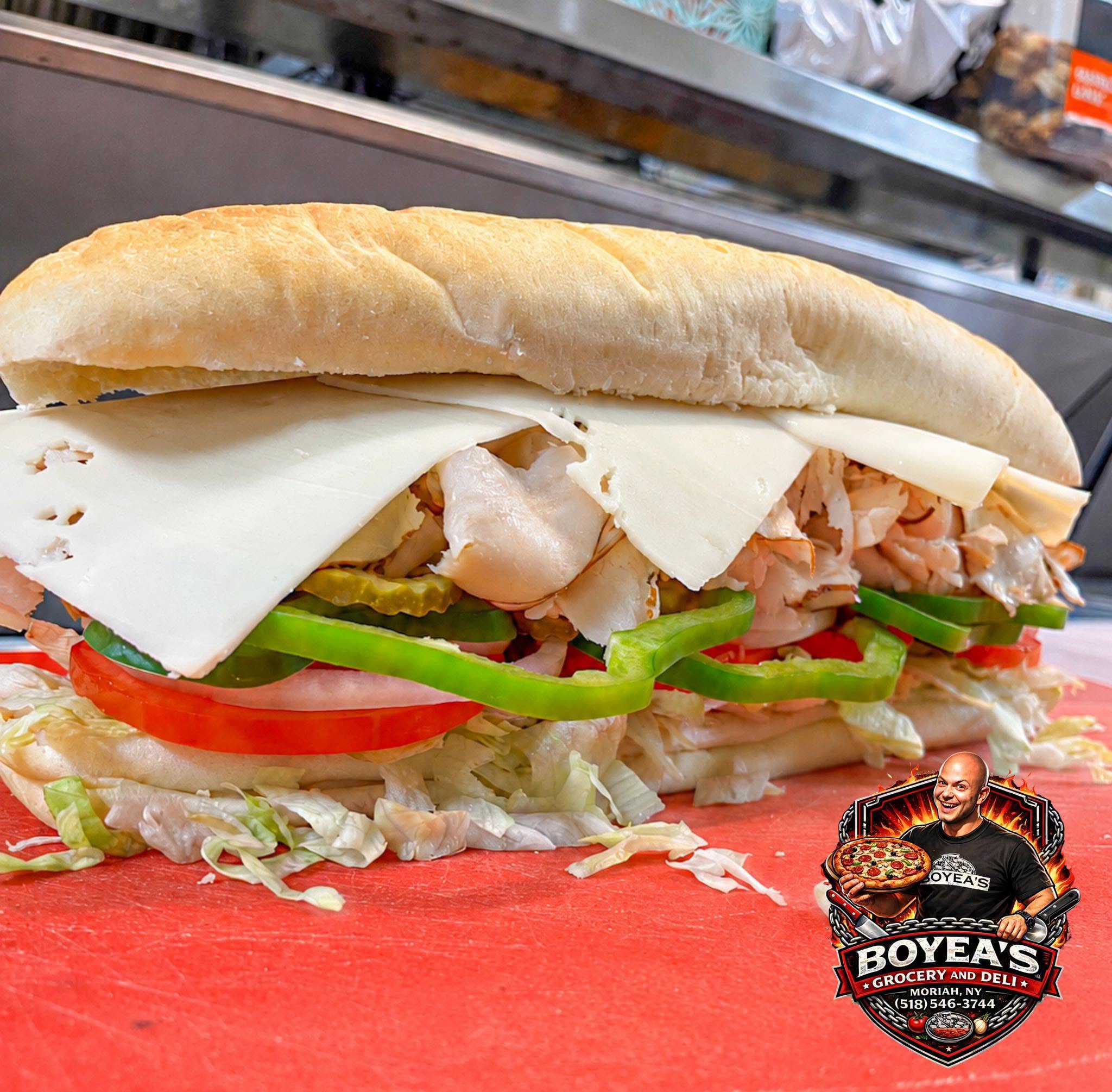 Close-up of a stacked Italian hoagie sandwich with various meats, cheeses, lettuce, tomato, cucumber, and pickles on a red surface in a grocery store.