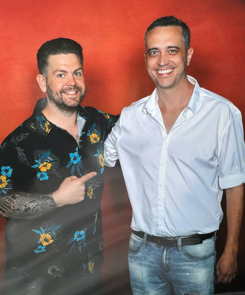 Two men smiling and standing close together against a red background, one pointing at the other.
