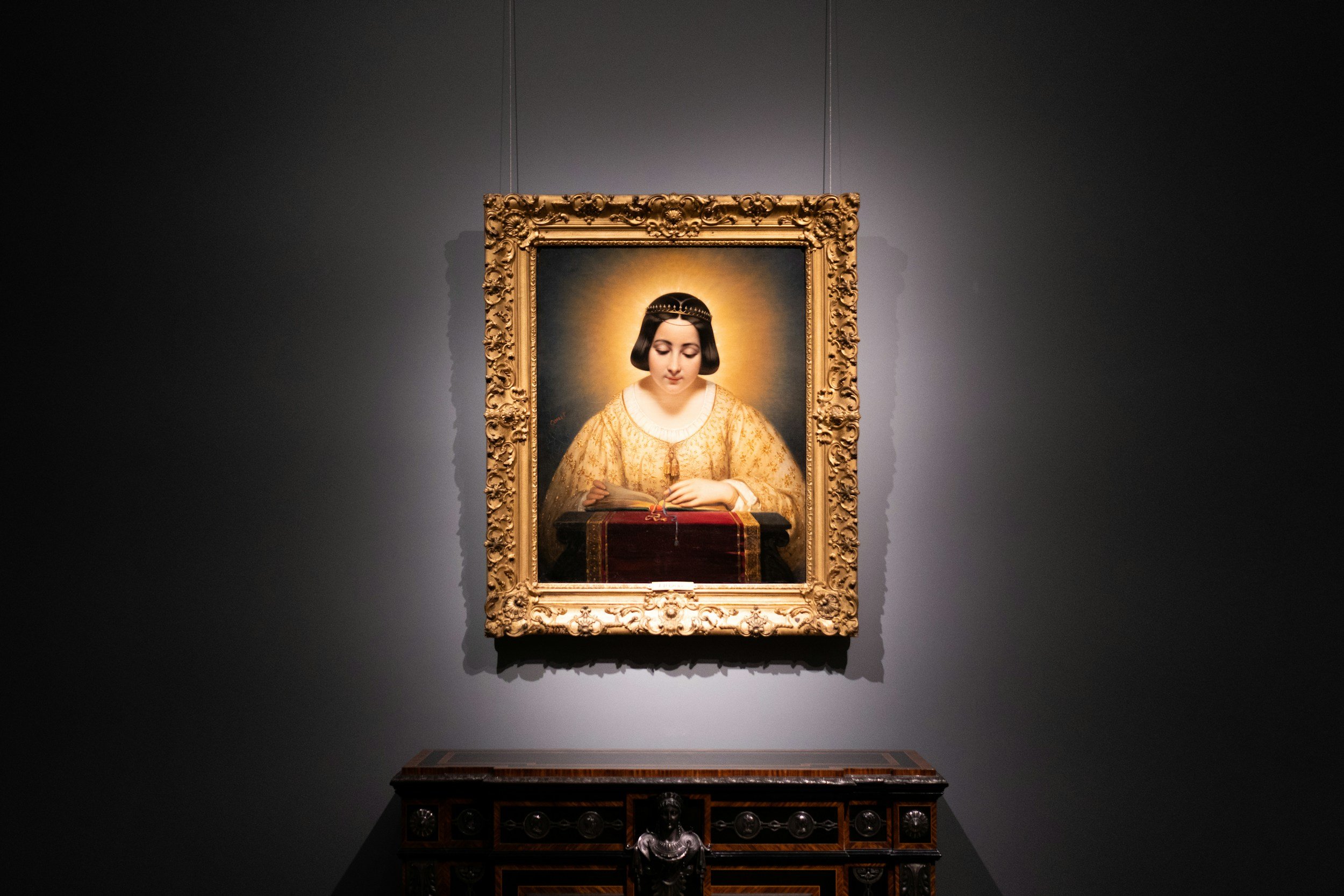 A portrait painting of a woman with a serene expression, wearing a golden dress with intricate patterns, and a jeweled headband, displayed on a dark gray wall in an art gallery.