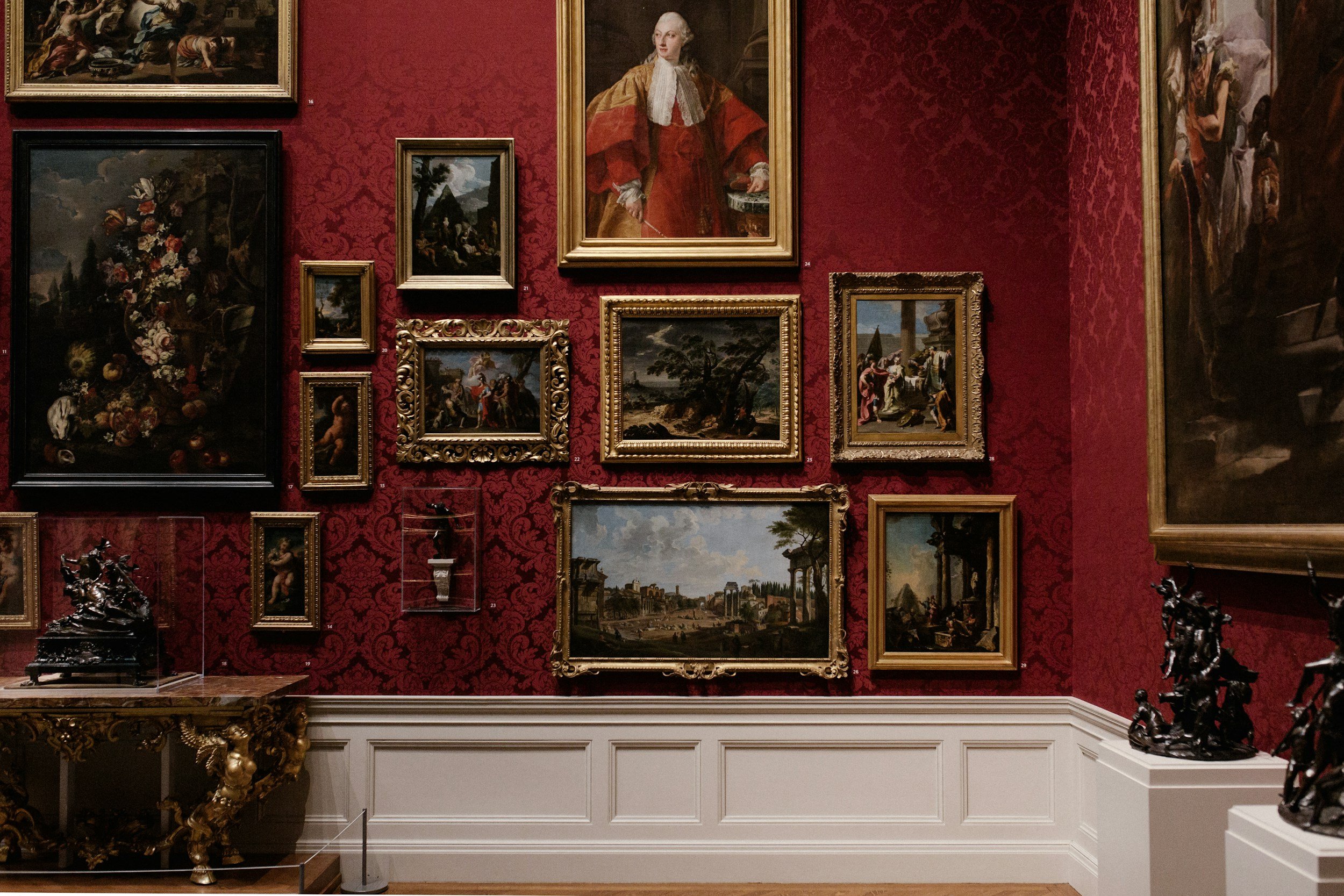 A gallery wall with various framed classical paintings on a red patterned wallpaper background. There are sculptures on pedestals and a table at the bottom with ornate decorative objects in a historic or art museum setting.