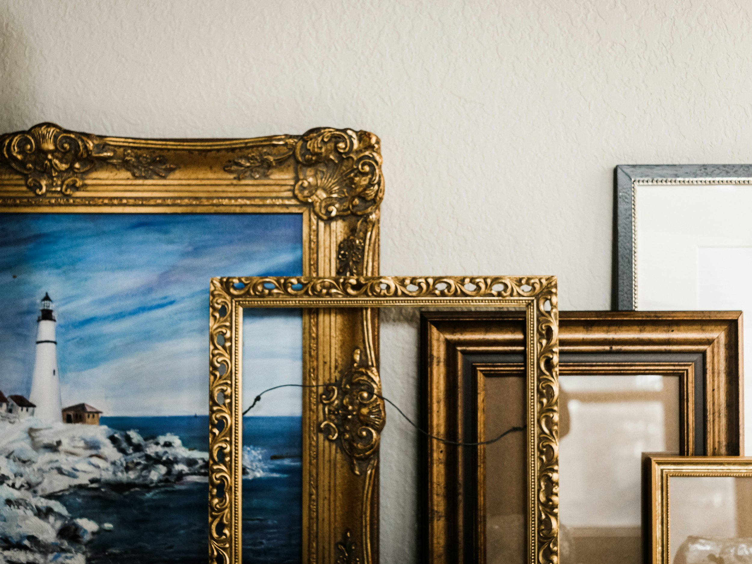Multiple ornate picture frames leaning against a light-colored textured wall, with a painting of a lighthouse and ocean in the background.