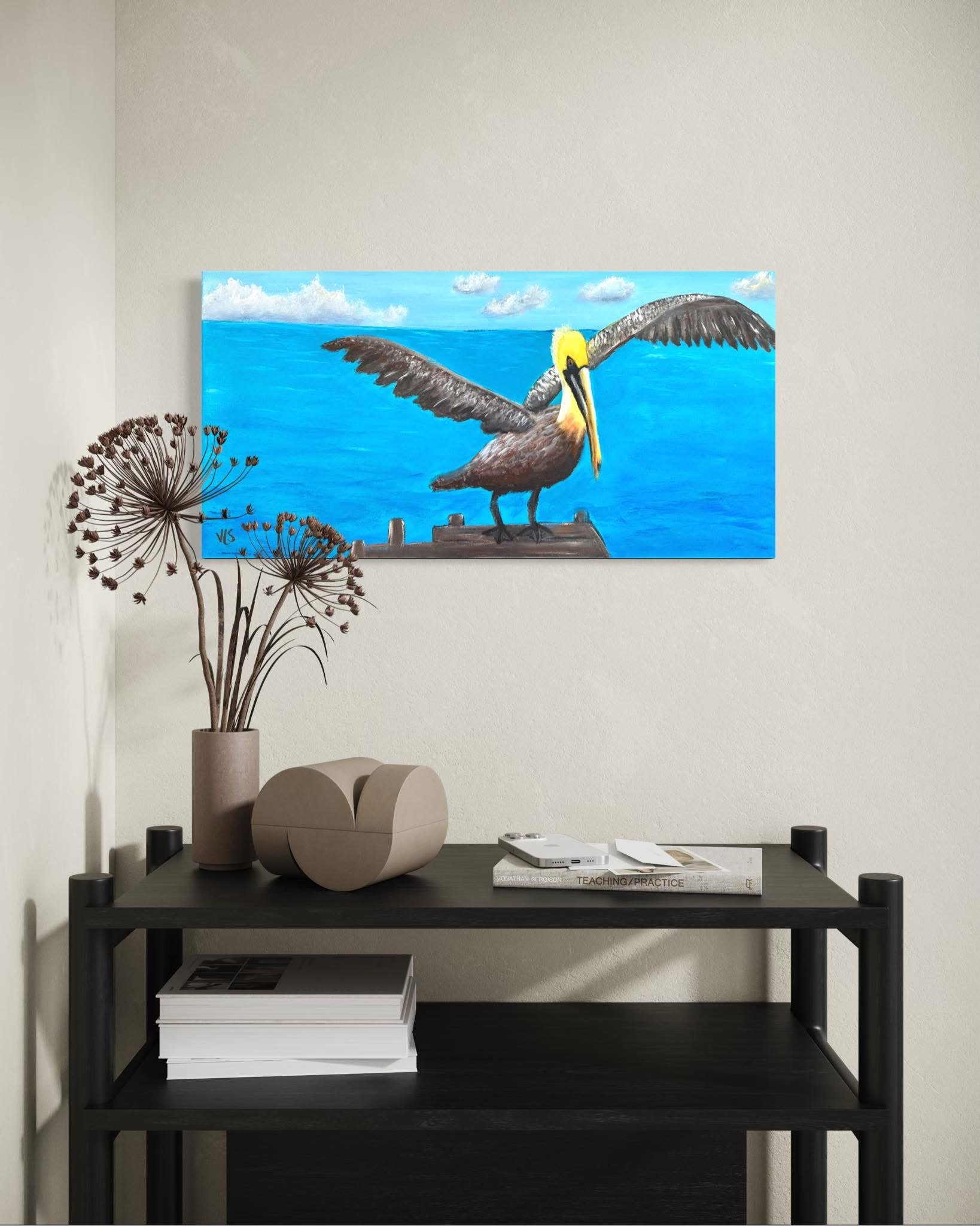 A black table with two tiers, a white vase with dried flowers, a stack of books, and a painting of a brown pelican on the wall behind it.