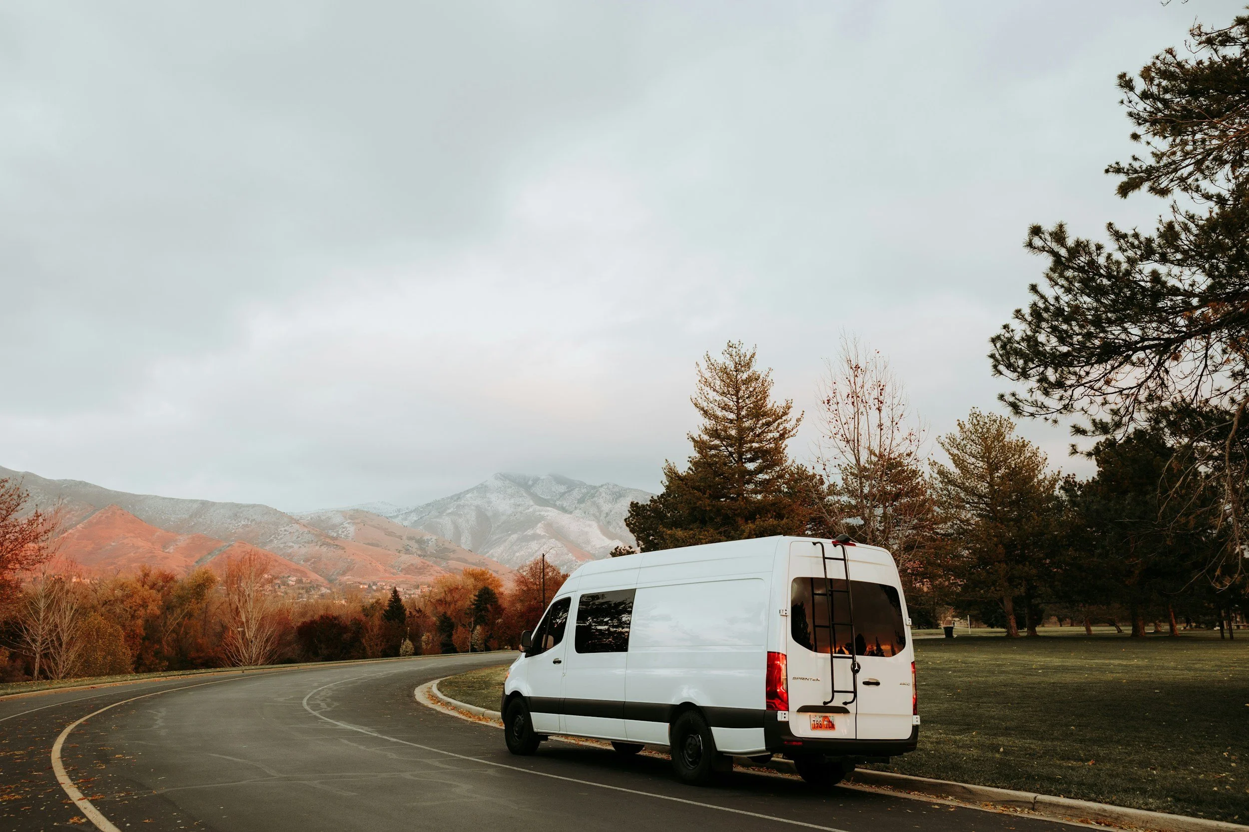 A white van parked on a curved road in a park with trees and mountains in the background during fall.