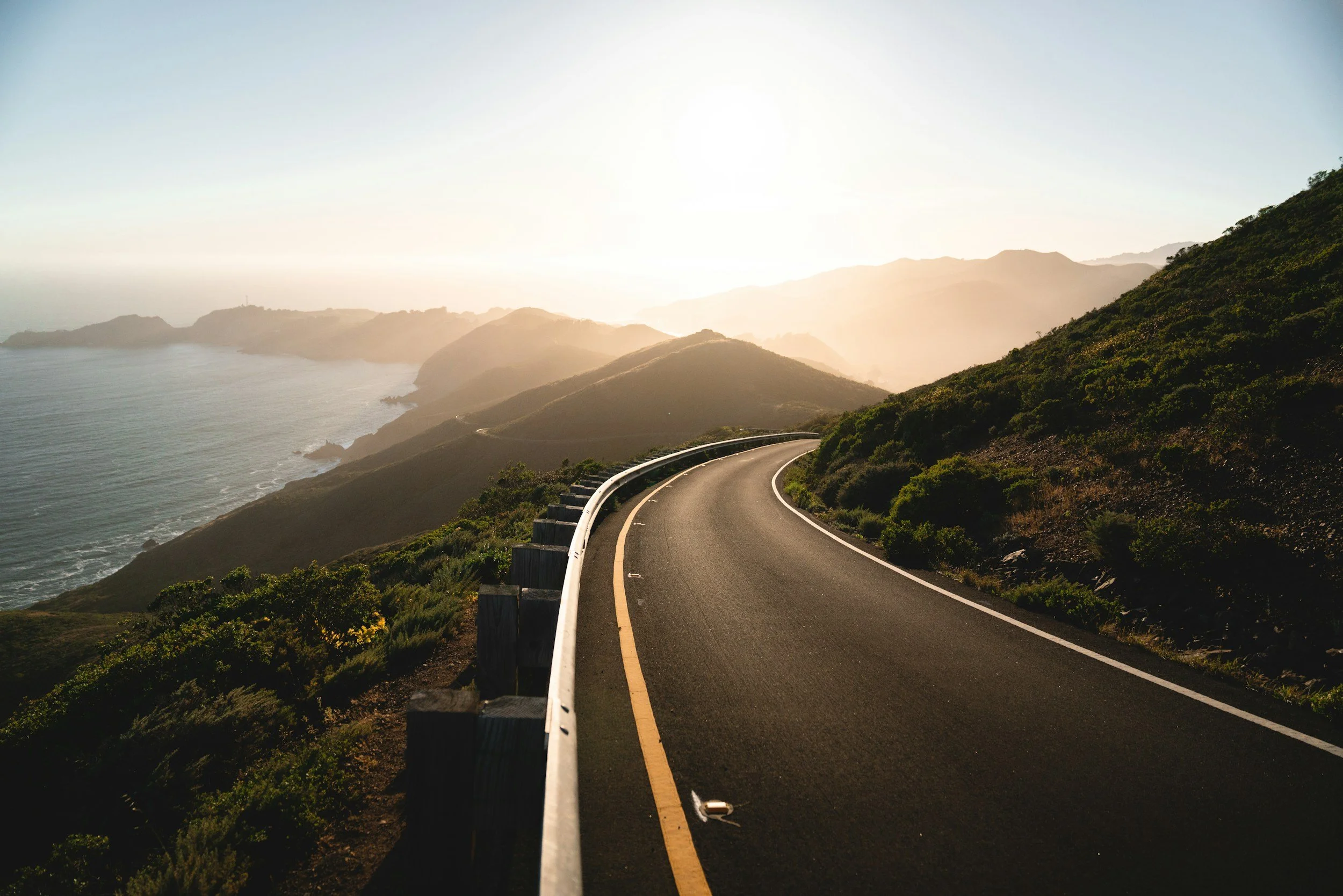 A winding coastal road with a mountain landscape on the right and the ocean on the left, under a bright sky.