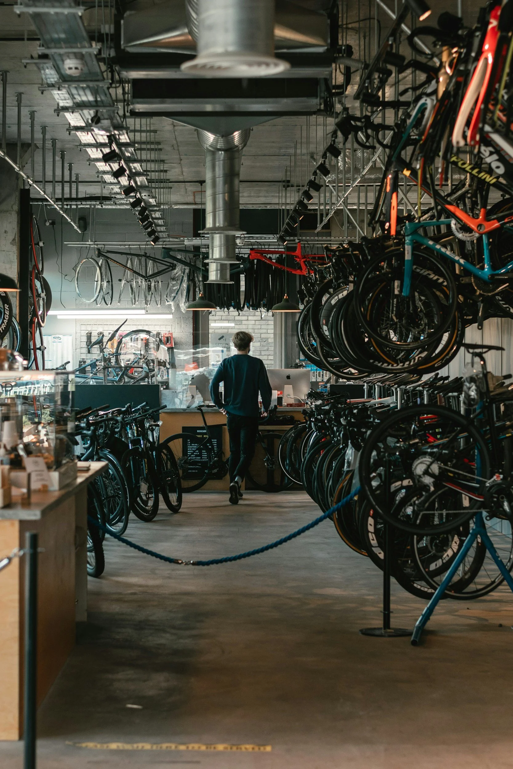 A person with short hair, wearing a dark shirt, stands inside a bicycle shop. The shop has bicycles hanging from the ceiling and displayed on the floor, with a counter and computer in the background. The environment has a modern industrial design with exposed concrete walls and ductwork.