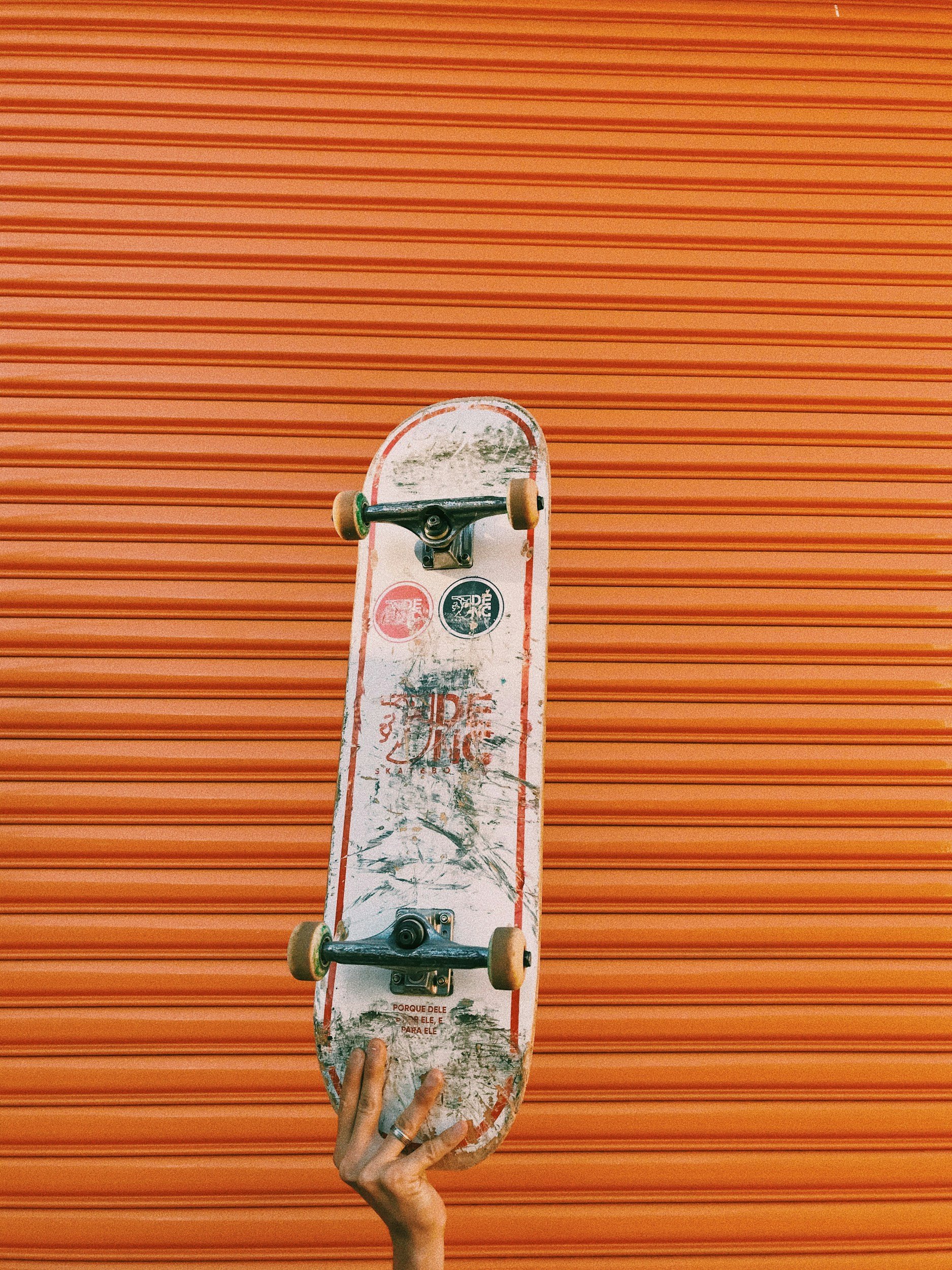Hand holding a skateboard in front of an orange corrugated metal wall.