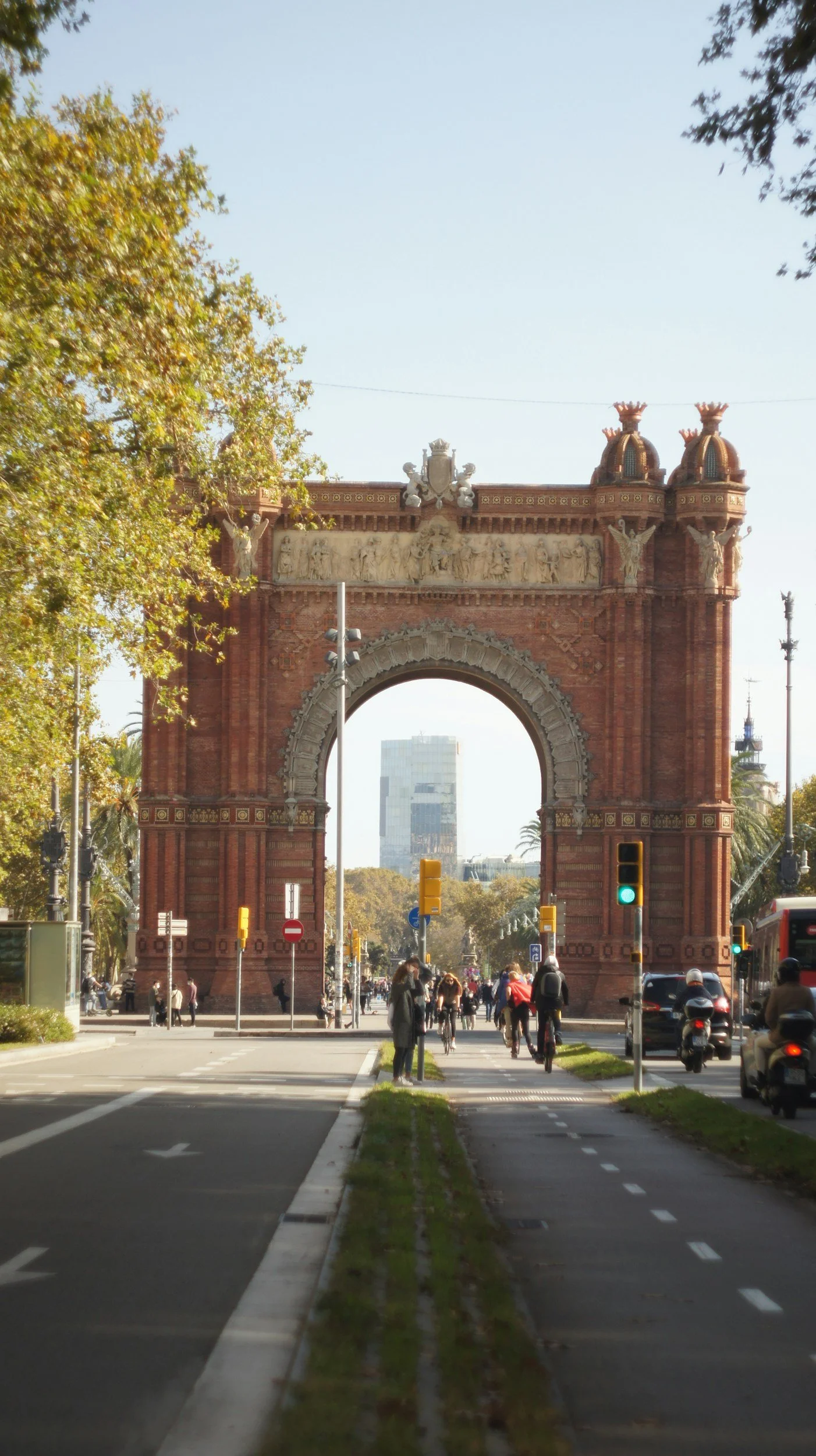 A historic archway in a city street with pedestrians, cyclists, and vehicles passing underneath, surrounded by trees and modern buildings in the background.