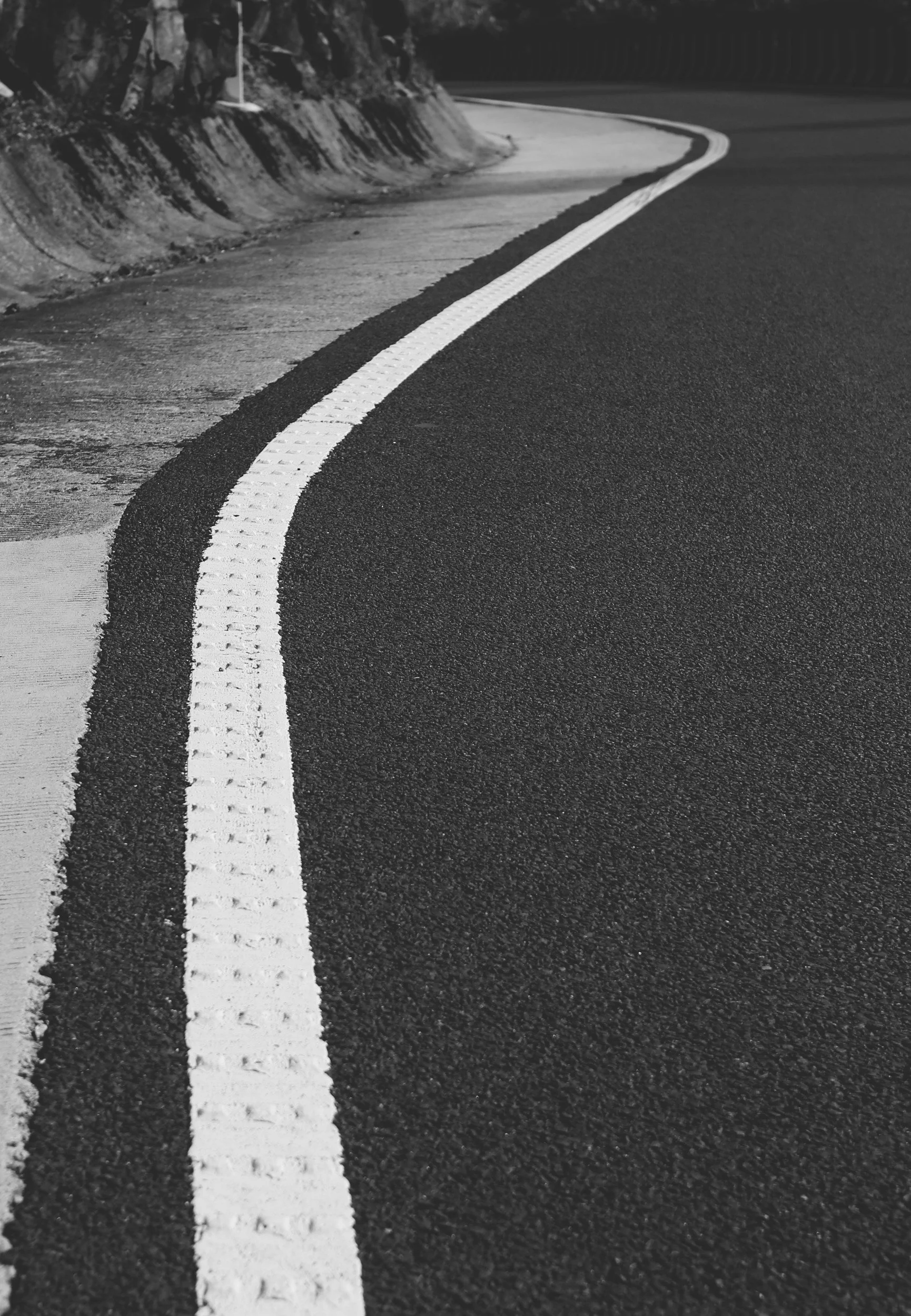 Black and white photograph of a curved road with a solid white line and a dotted white line, alongside a rocky hillside.