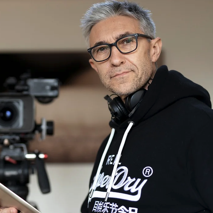 A man with gray hair, wearing glasses and a black hoodie with white lettering, standing near a professional camera on a tripod.