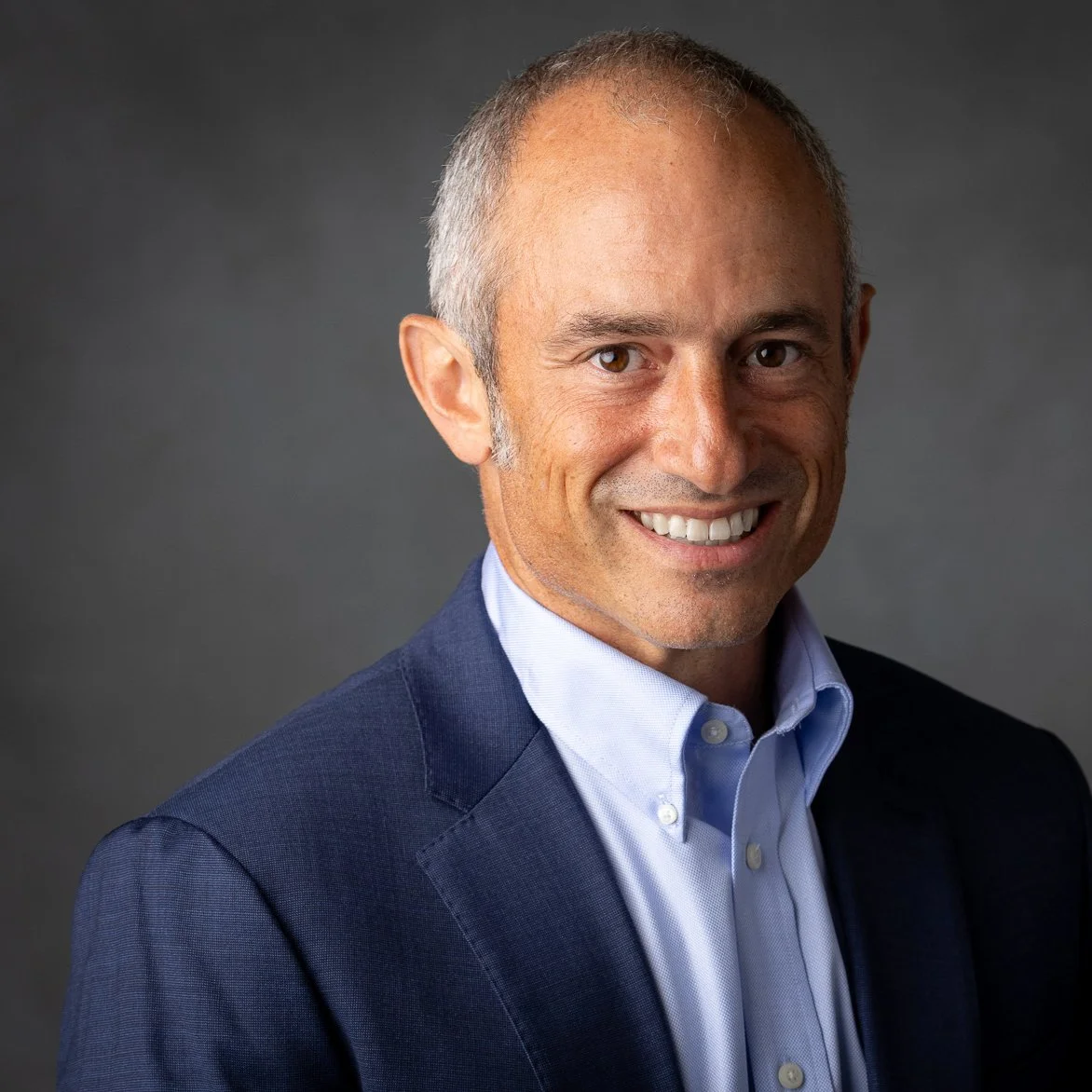 Portrait of a smiling man with short gray hair, wearing a blue suit jacket and light blue collared shirt, against a dark gray background.