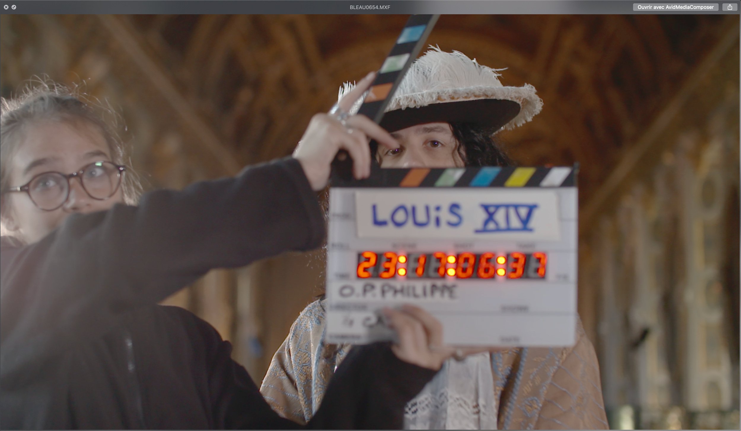 A person holding a clapperboard marked 'Louis XIV' with a man in historical costume wearing a feathered hat beside them, on a film set with a cathedral interior background.