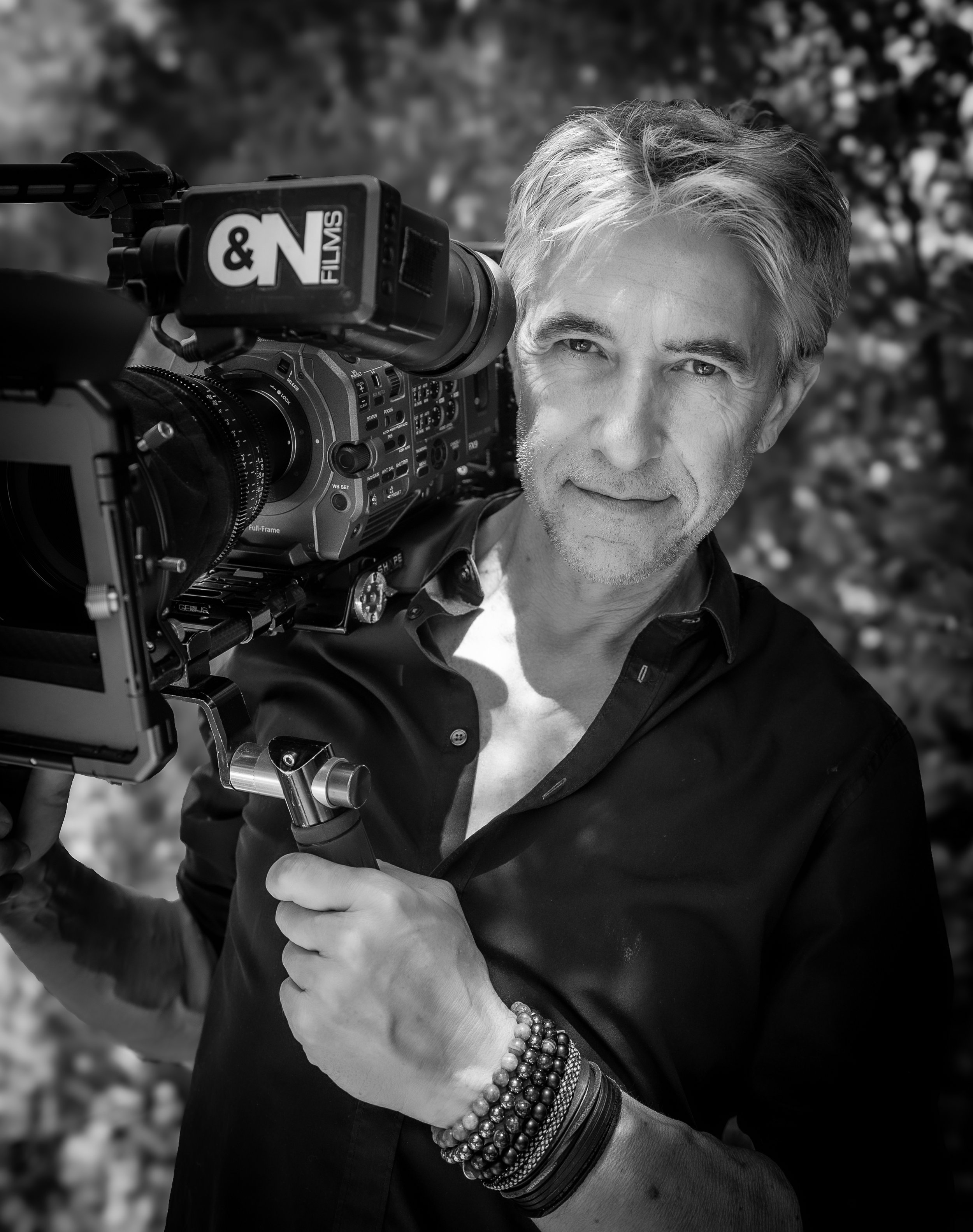 Black and white photo of a Oliver Paul Philippe holding a professional video camera with an 'O&N Films' logo while smiling and looking at the camera.