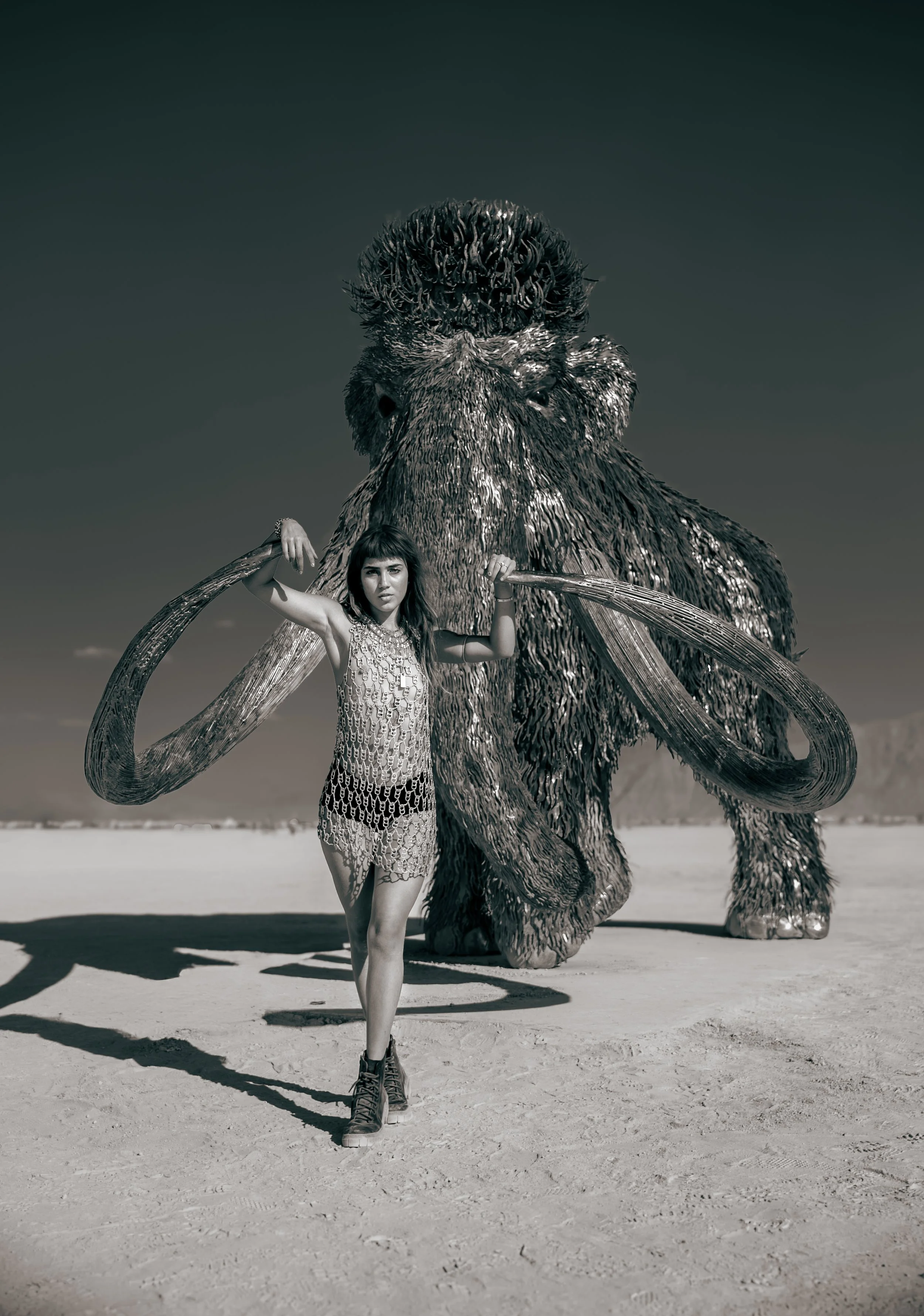A woman in a dress and combat boots standing on the Burning Man Playa desert with a large, furry mammoth sculpture in the background.