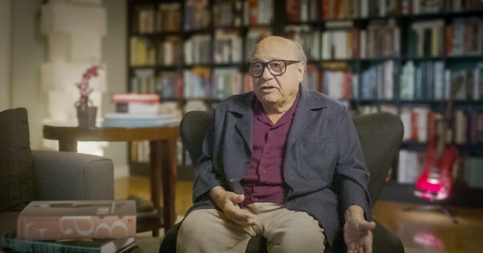 A photo of Danny DeVito. He is wearing glasses, a maroon shirt, and a dark blazer sits in a living room with a large bookshelf background, engaged in conversation.