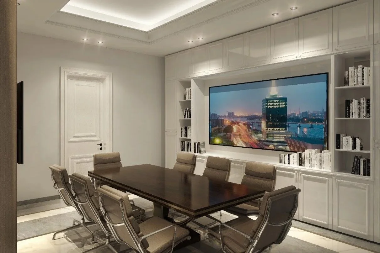Conference room with a large wooden table, eight beige chairs, white built-in shelving with books, and a large monitor displaying a cityscape at night.