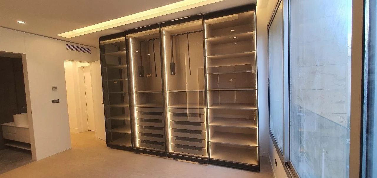 A room with a large glass-fronted display cabinet or wine cooler with shelves, illuminated by internal lighting, next to a large window letting in natural light.