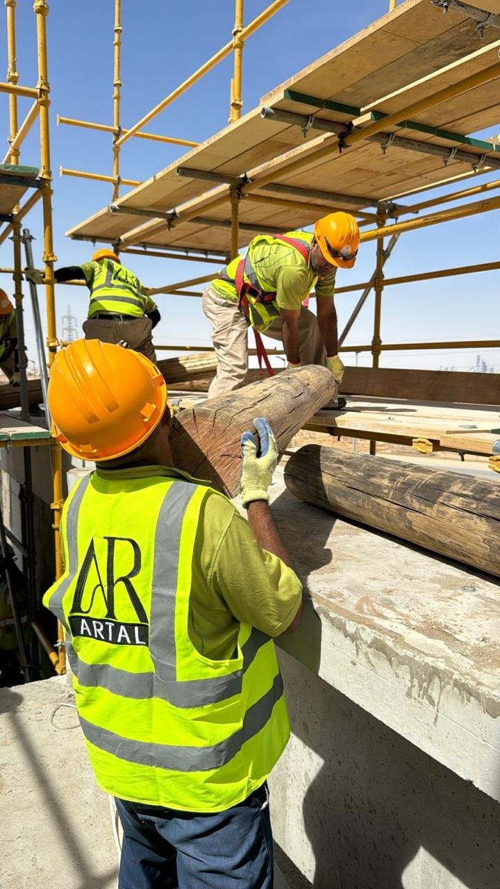 Construction workers in safety vests and yellow helmets installing large wooden beams on a scaffolding structure at a construction site.