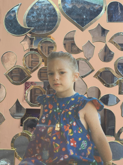 A young girl with blonde hair tied back, wearing a colorful dress with a pattern of geometric shapes, sitting in front of a decorative wall with multiple mirror frames of various shapes and sizes. The mirror frames are gold-colored and reflect parts of the room behind the camera.