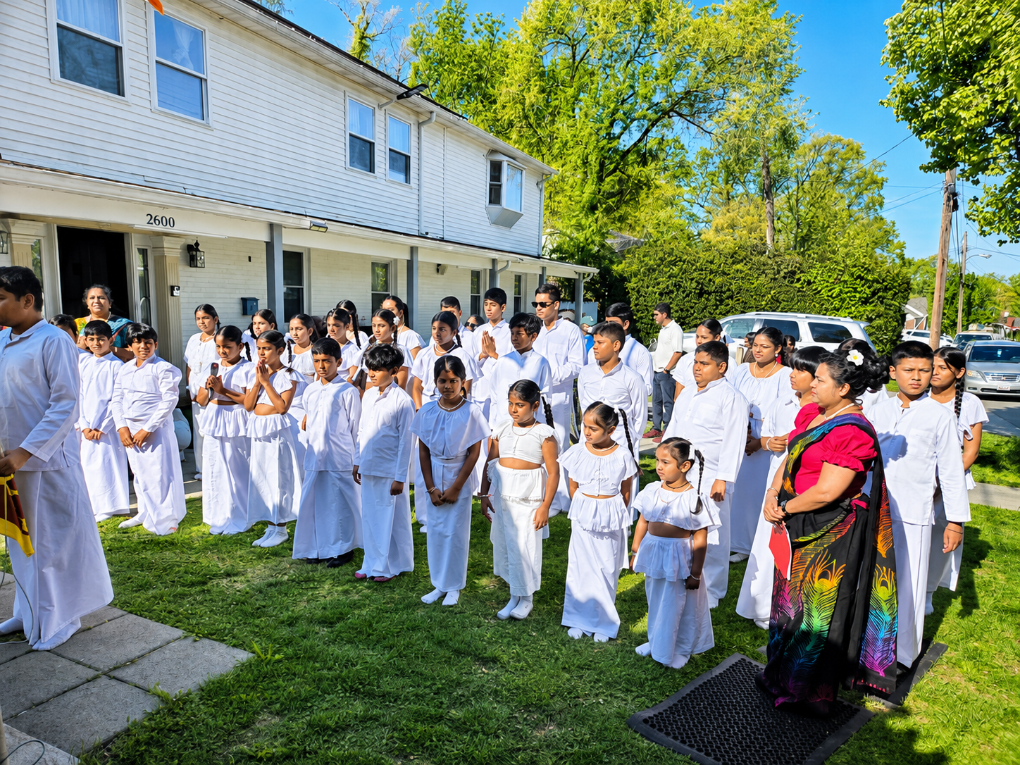 Group of people, mostly children dressed in white outfits, standing outside for a ceremonial or religious activity in front of a building with a white facade and a lawn, on a sunny day.