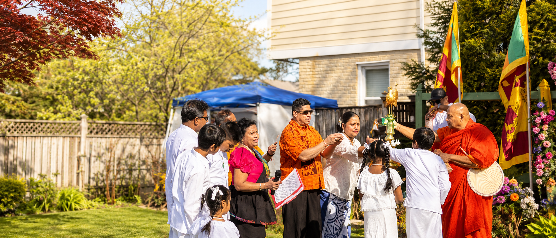 A group of people participating in a religious or cultural ceremony outdoors, with a Buddhist monk in orange robes, Sri Lankan flags, and floral decorations, on a sunny day in a backyard with trees and a house in the background.