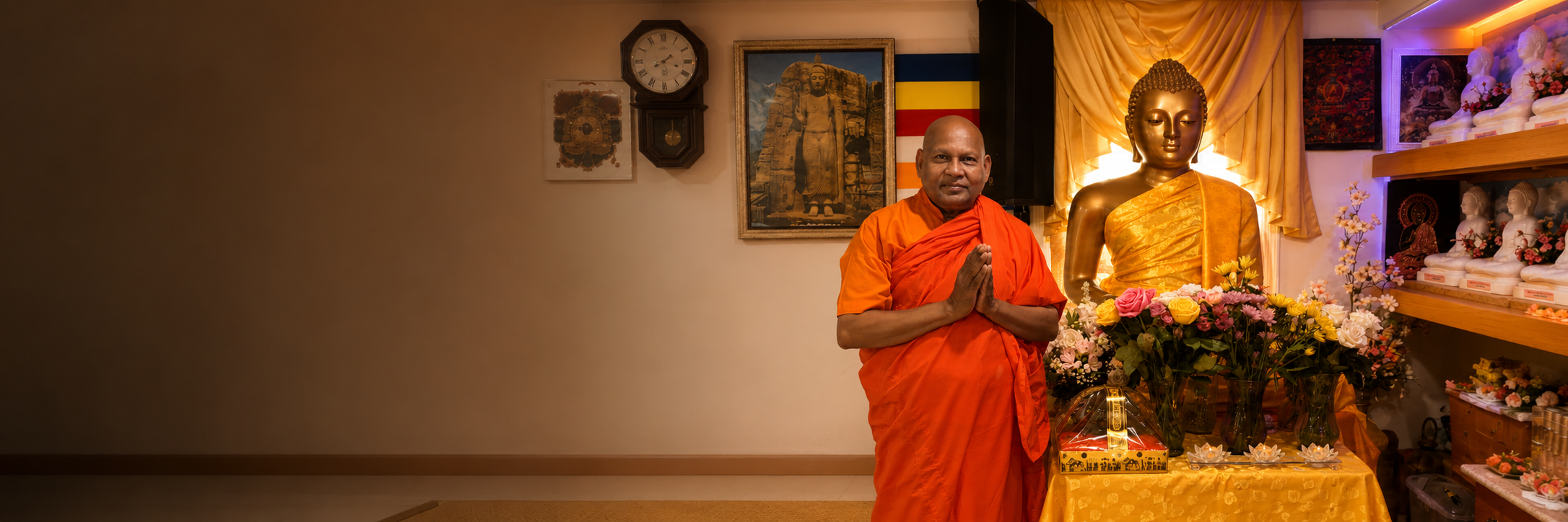 A man in orange robes standing in prayer with hands pressed together in front of a large golden Buddha statue surrounded by flowers in a shrine or temple setting.