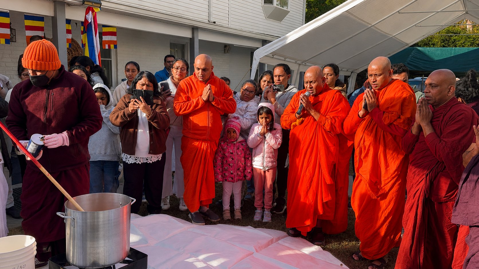 Group of people, including monks in orange robes, gathered outdoors under a white canopy, praying and participating in a religious ceremony. Some hold prayer beads and smartphones.