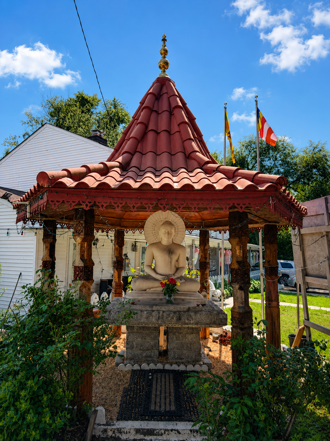 A white Buddha statue sitting cross-legged under a red-tiled, pointed roof shrine with wooden columns, located outside on a bright sunny day with blue sky, white clouds, and American flags in the background.