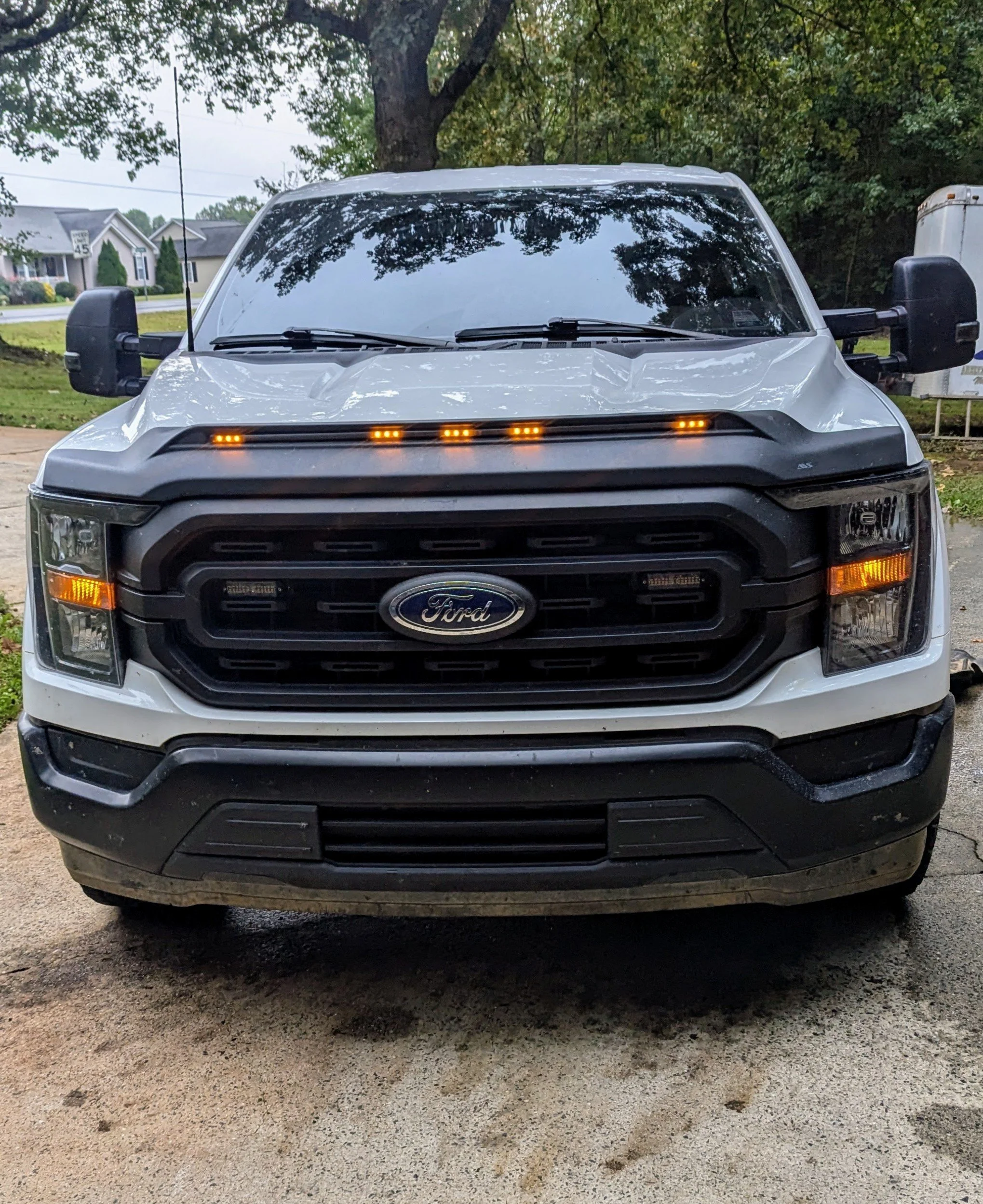 Front view of a white Ford truck parked on dirt ground with trees and houses in the background.