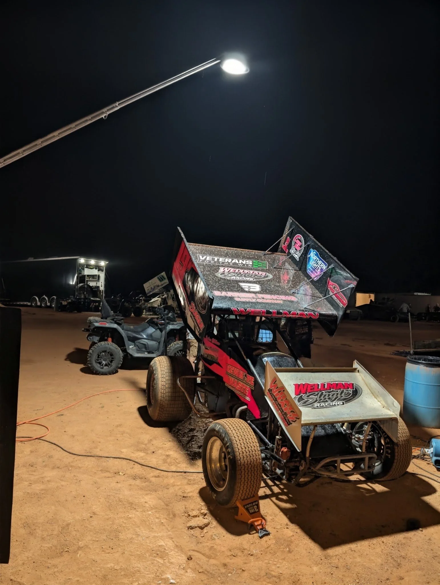 A racing go-kart with sponsorship decals on a dirt track at night, with a race track and equipment visible in the background under bright lights.