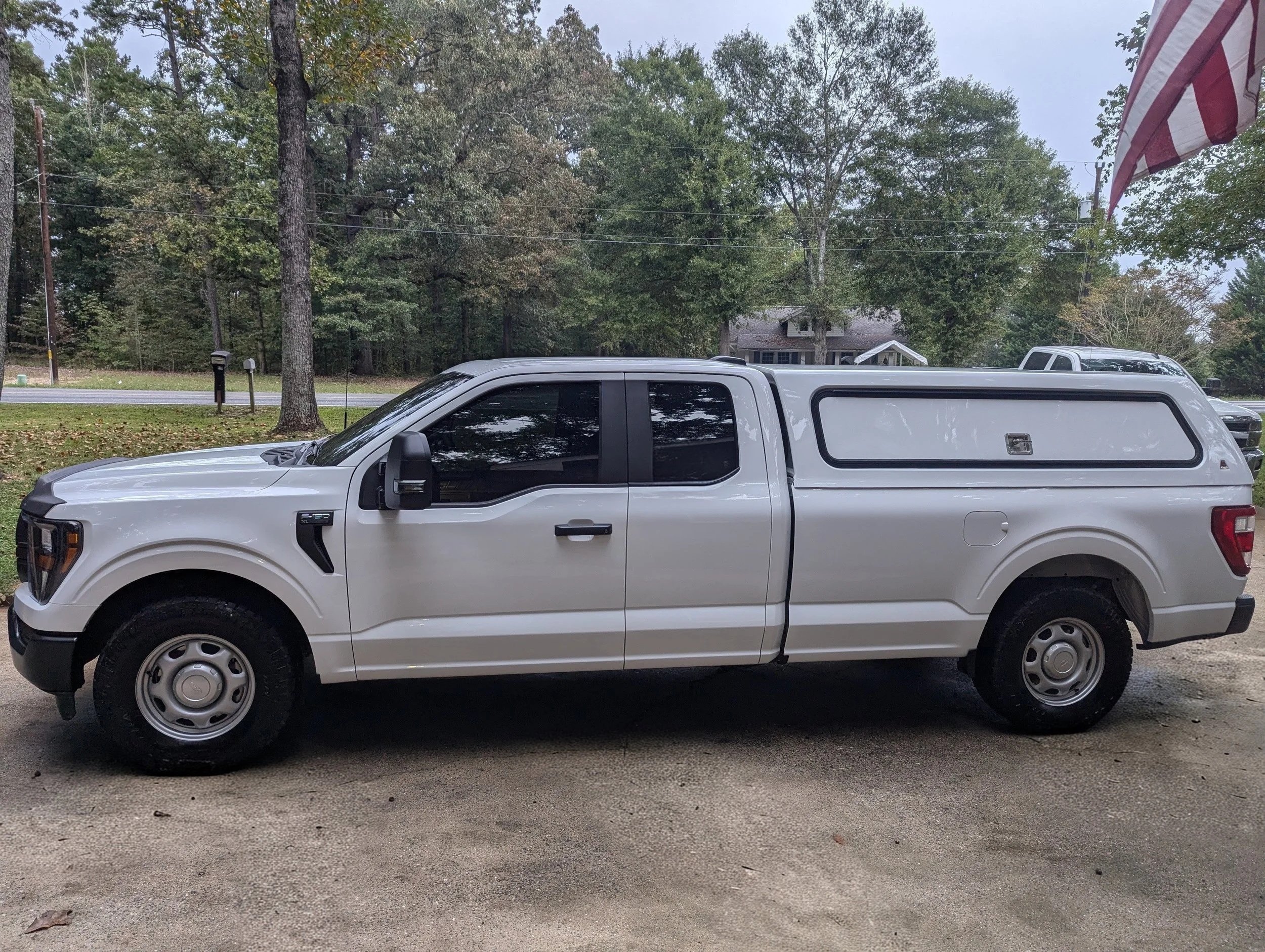 A white pickup truck parked outdoors in a driveway or parking area, with trees and a house in the background, and part of an American flag visible on the right side of the image.