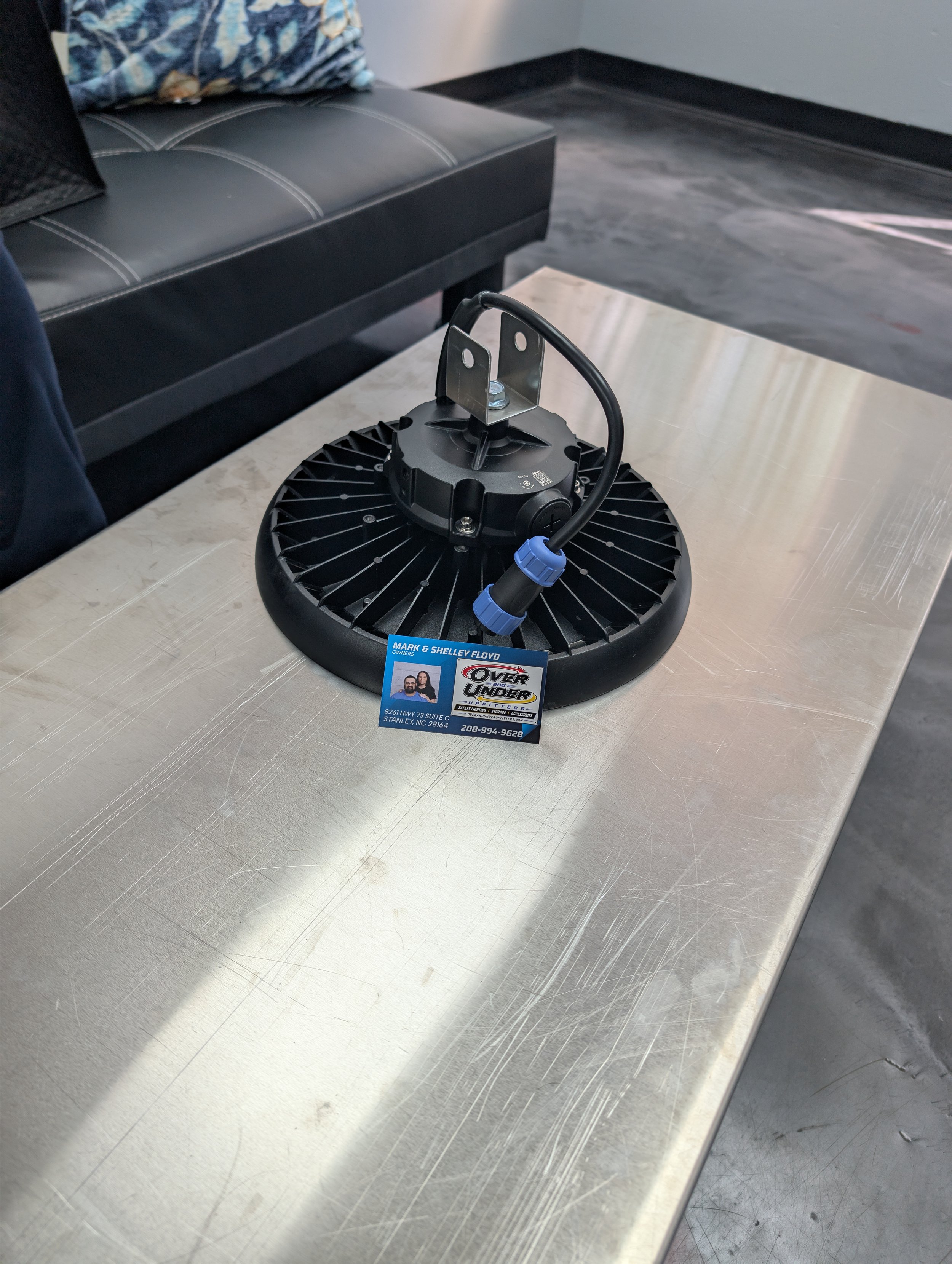 A metallic device with a black circular base, a central mounting bracket, and a black cable with blue connectors placed on a stainless steel table. In the background, a black leather bench with white stitching and a colorful pillow, along with a gray wall and floor.