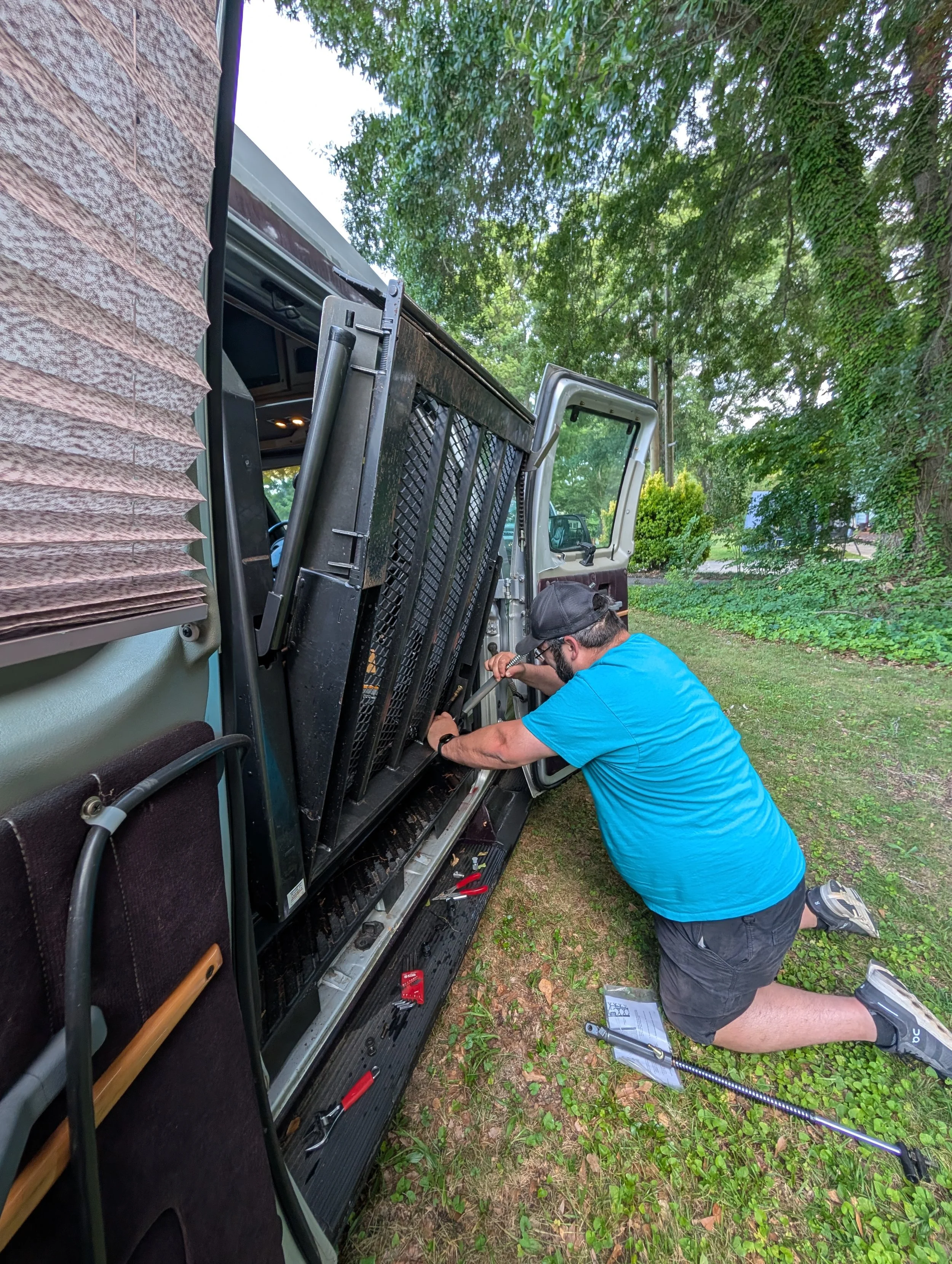 A man kneeling outside next to a black truck, working on a vehicle part with tools, surrounded by green trees and grass.