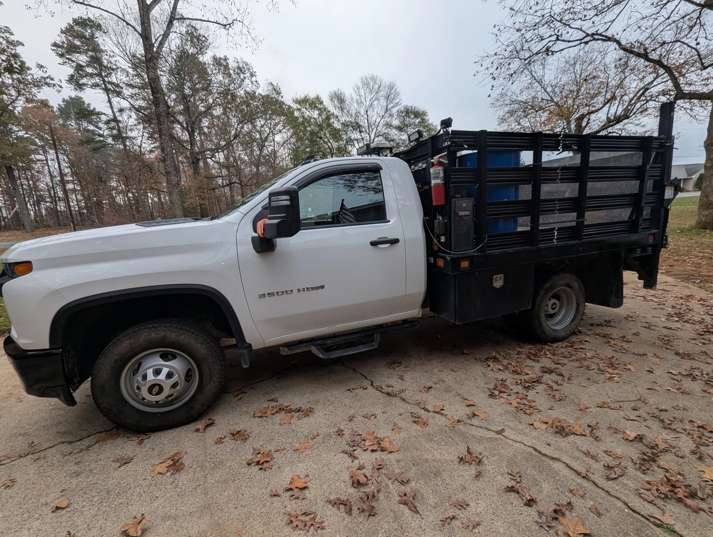 A white pickup truck with a black flatbed carrying blue barrels and equipment, parked on a street with fallen autumn leaves, trees with sparse branches in the background.