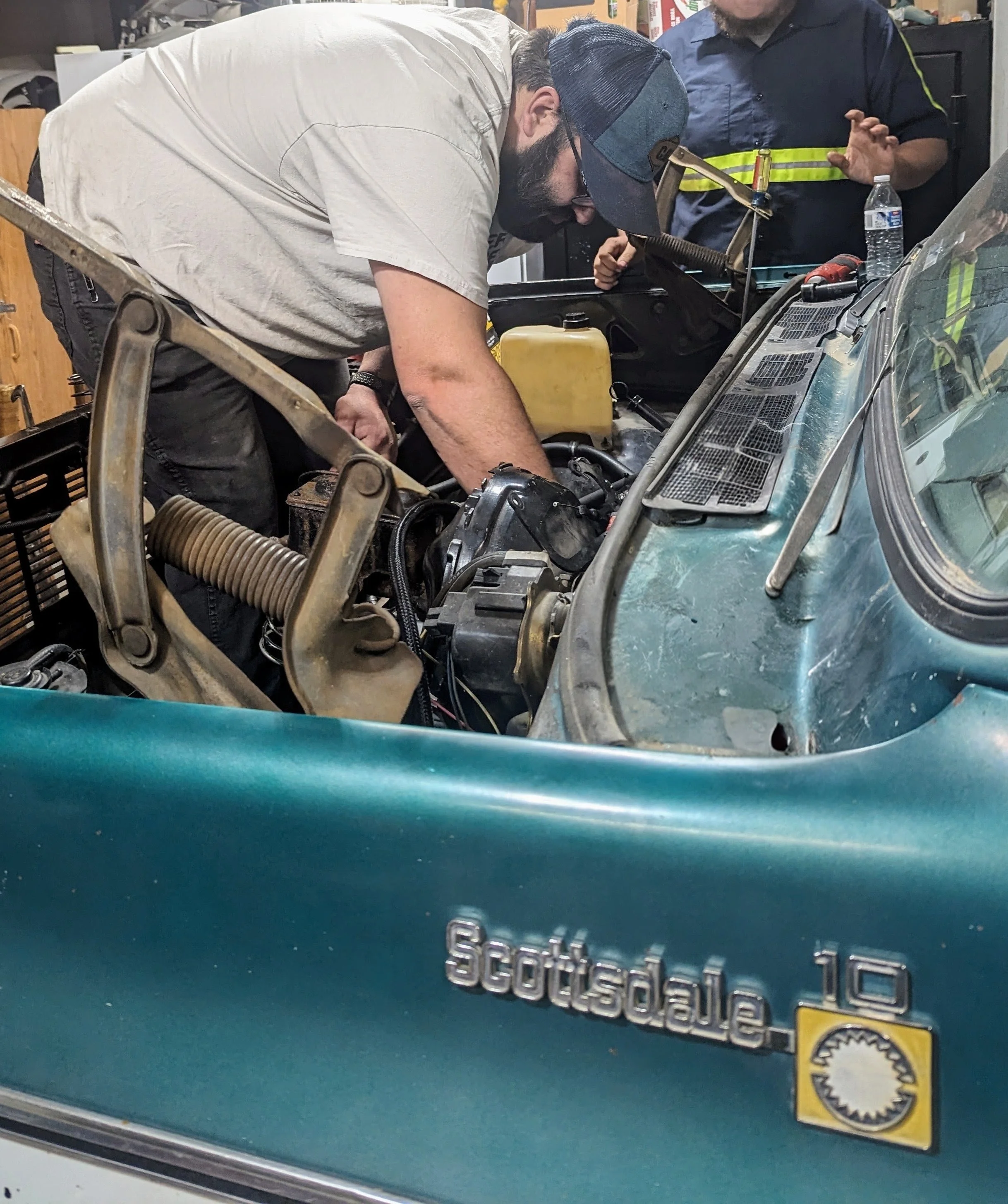 Man working on a car engine in a garage, with a woman nearby observing. The car has a windshield with a sticker that reads 'Scuderia.'