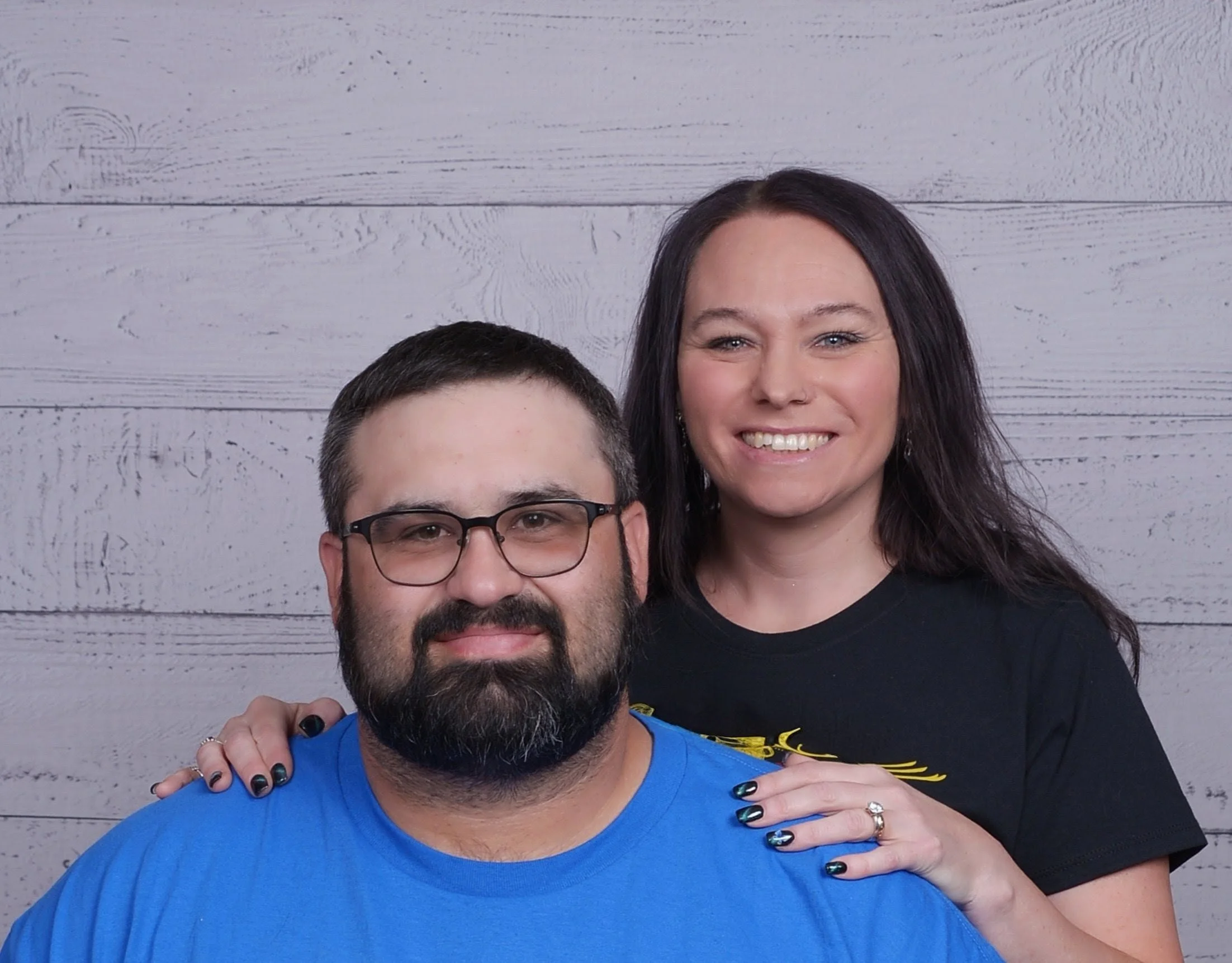 A woman and a man pose for a photo against a white wooden background. The woman has long dark hair, is smiling, and is wearing a black shirt with yellow accents. The man has short dark hair, is wearing glasses and a blue shirt, and is smiling. The woman has her hand on the man's shoulder.