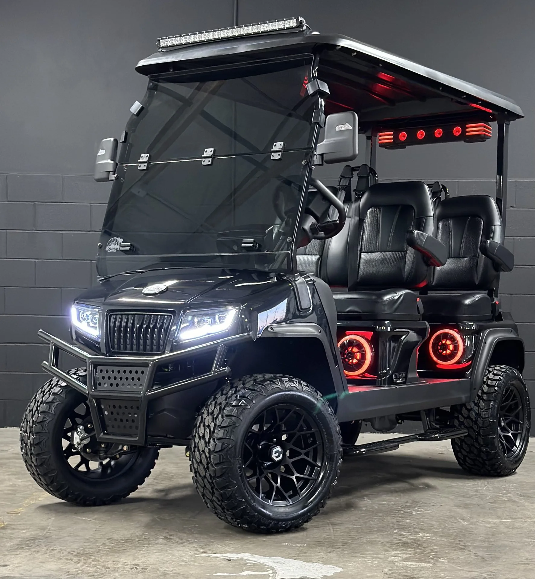 A black golf cart with custom wheels and tires, LED headlight, and additional lighting on the roof against a gray brick wall background.