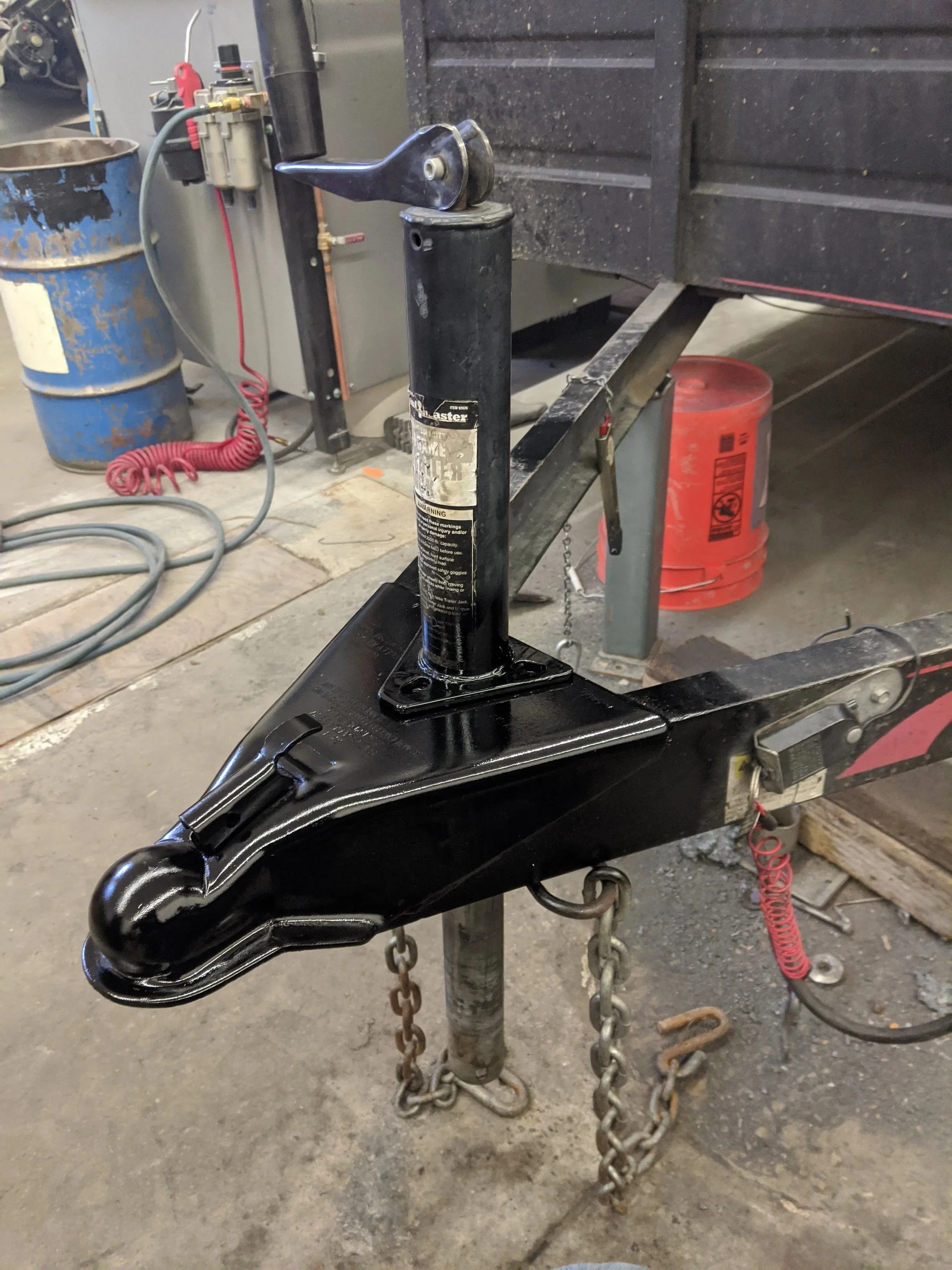 A black trailer hitch mounted on a metal pole, with chains hanging from it, inside a workshop or garage with tools, a barrel, and equipment in the background.