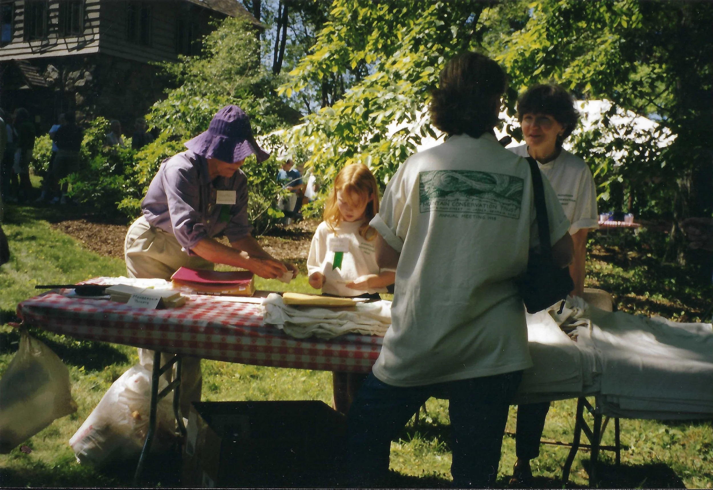 People gathered outdoors at a table with books and papers, surrounded by trees and greenery, during a sunny day.