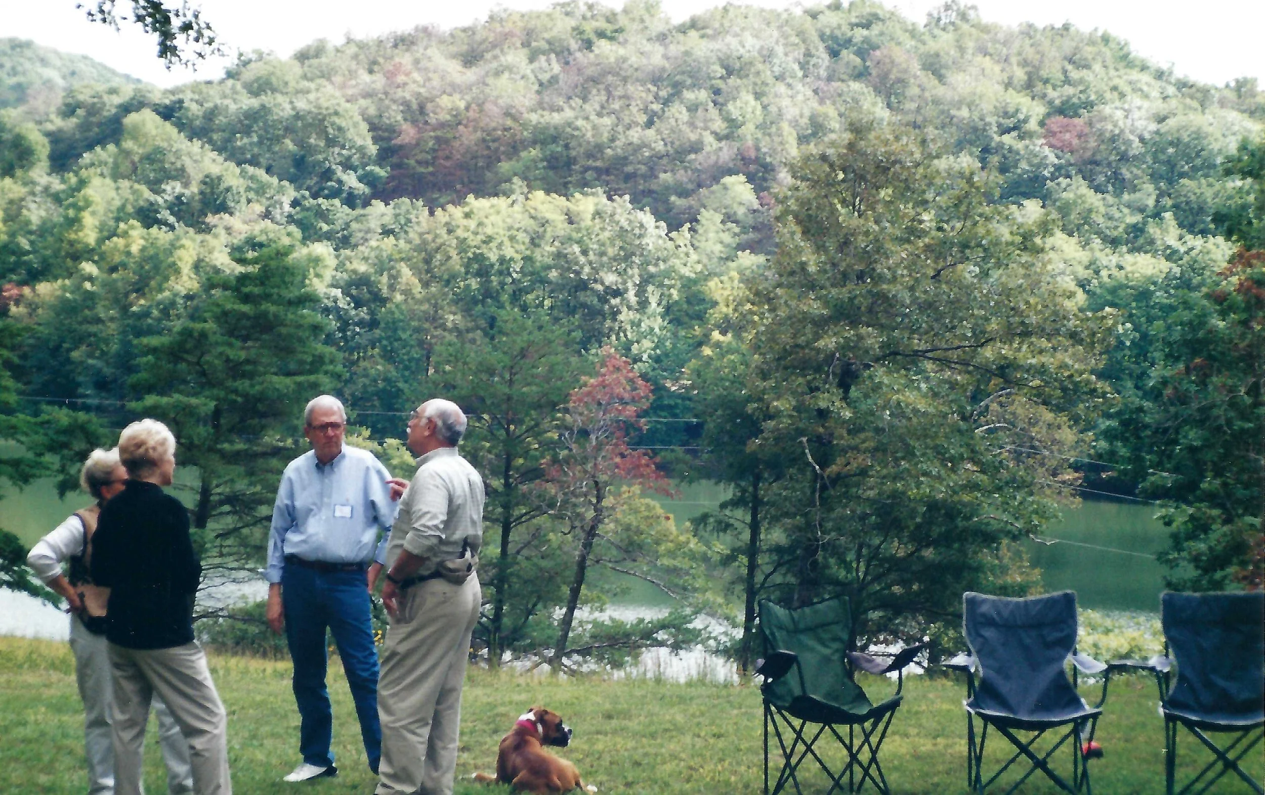 Group of five people standing near a lake, surrounded by trees, with two empty folding chairs and a dog sitting on the grass.