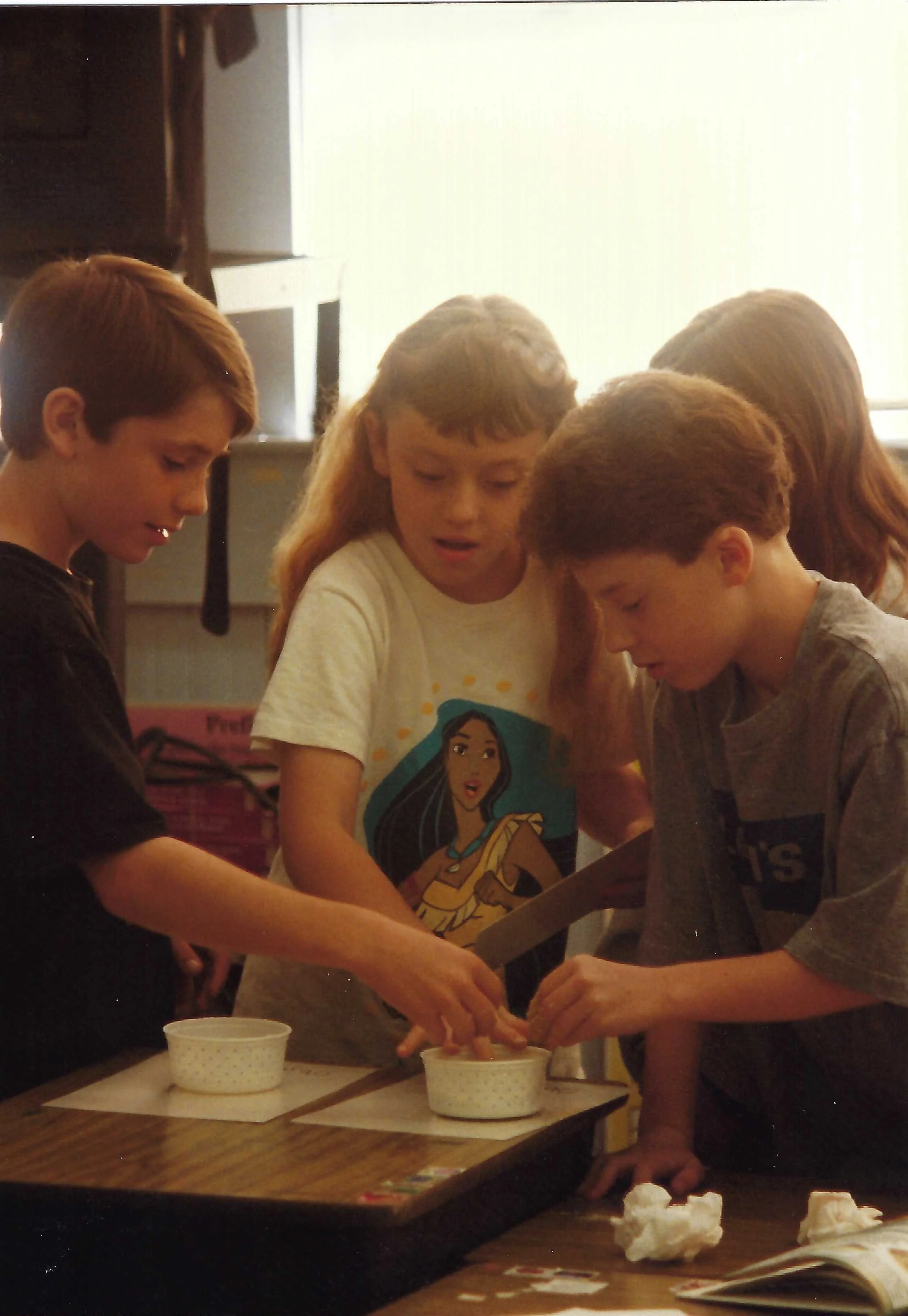 Four children gathered around a table playing a game involving tissue paper and cups in a classroom or game room.